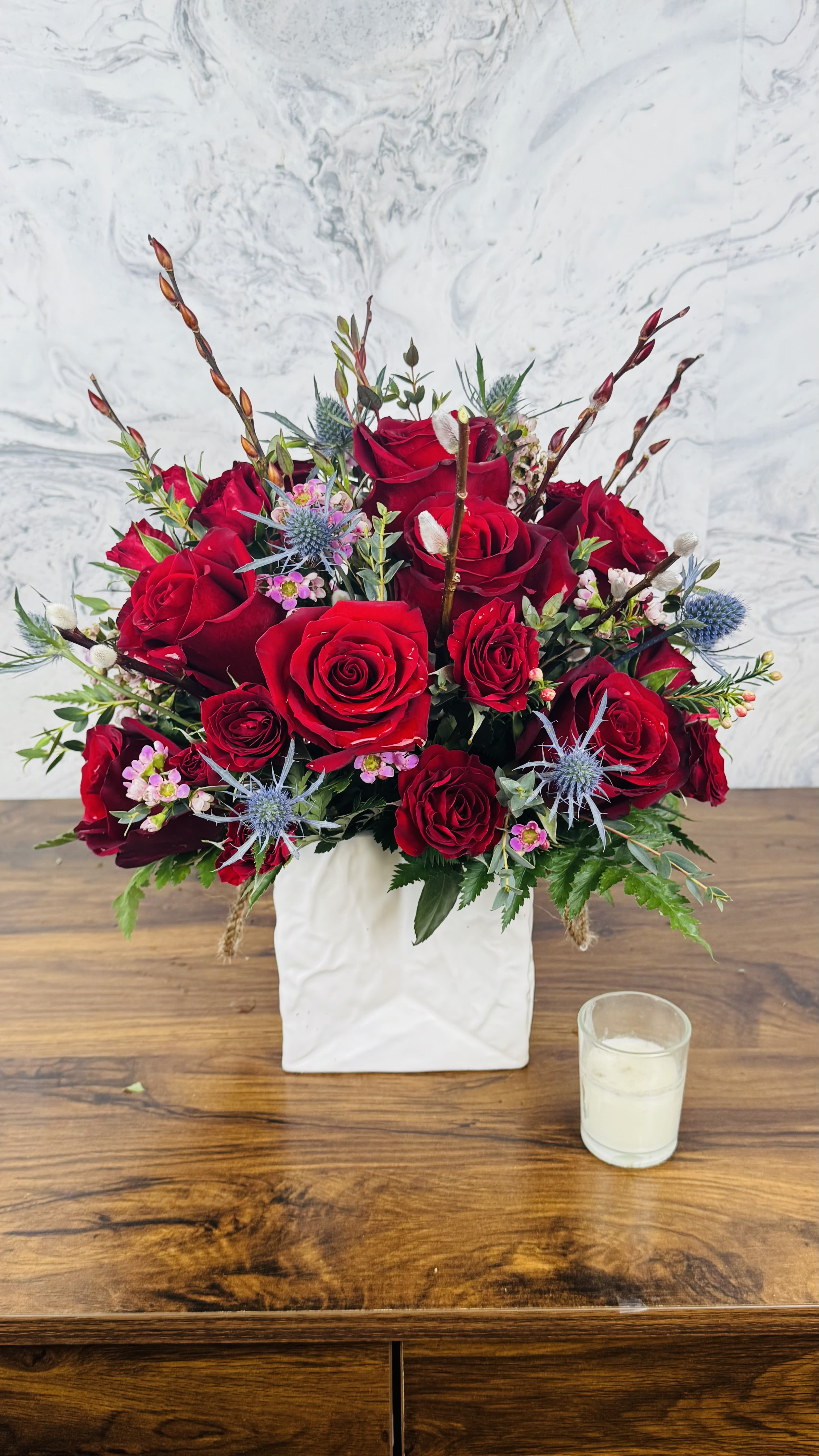 Red rose arrangement in a white square vase with blue thistle accents