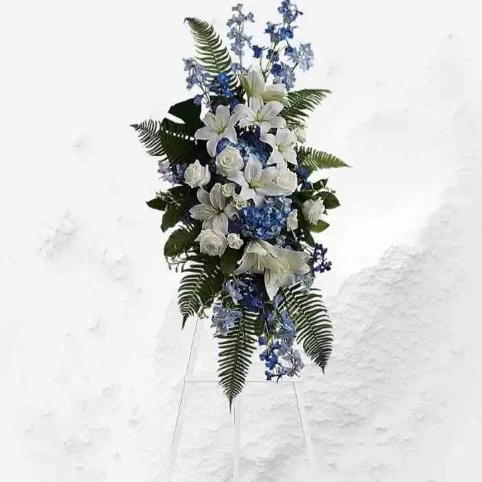 Ocean Breeze Spray - A tall standing spray in cool whites and ocean blues, perfect as a sympathy arrangement or a calm tribute for a memorial. White lilies and roses take center stage, their open blooms offering a gentle, respectful presence, while blue hydrangeas and delphinium add soft, soothing color that reads as peaceful and composed. The overall look is dignified and serene-the kind of floral expression that helps the person you're honoring feel remembered and respected.  Arranged on a white metal easel in an elongated, slightly cascading oval, the composition is framed with bold fern fronds that give height and a graceful vertical line. The mix of textures-from the structured roses and lilies to the cloud-like hydrangea clusters and spiky delphinium-creates visual depth while staying understated. Sized to stand prominently at a service or beside a memorial, this spray arrives ready to display on its included white metal easel and is designed to convey quiet sympathy and lasting regard.funeral and sympathy spray