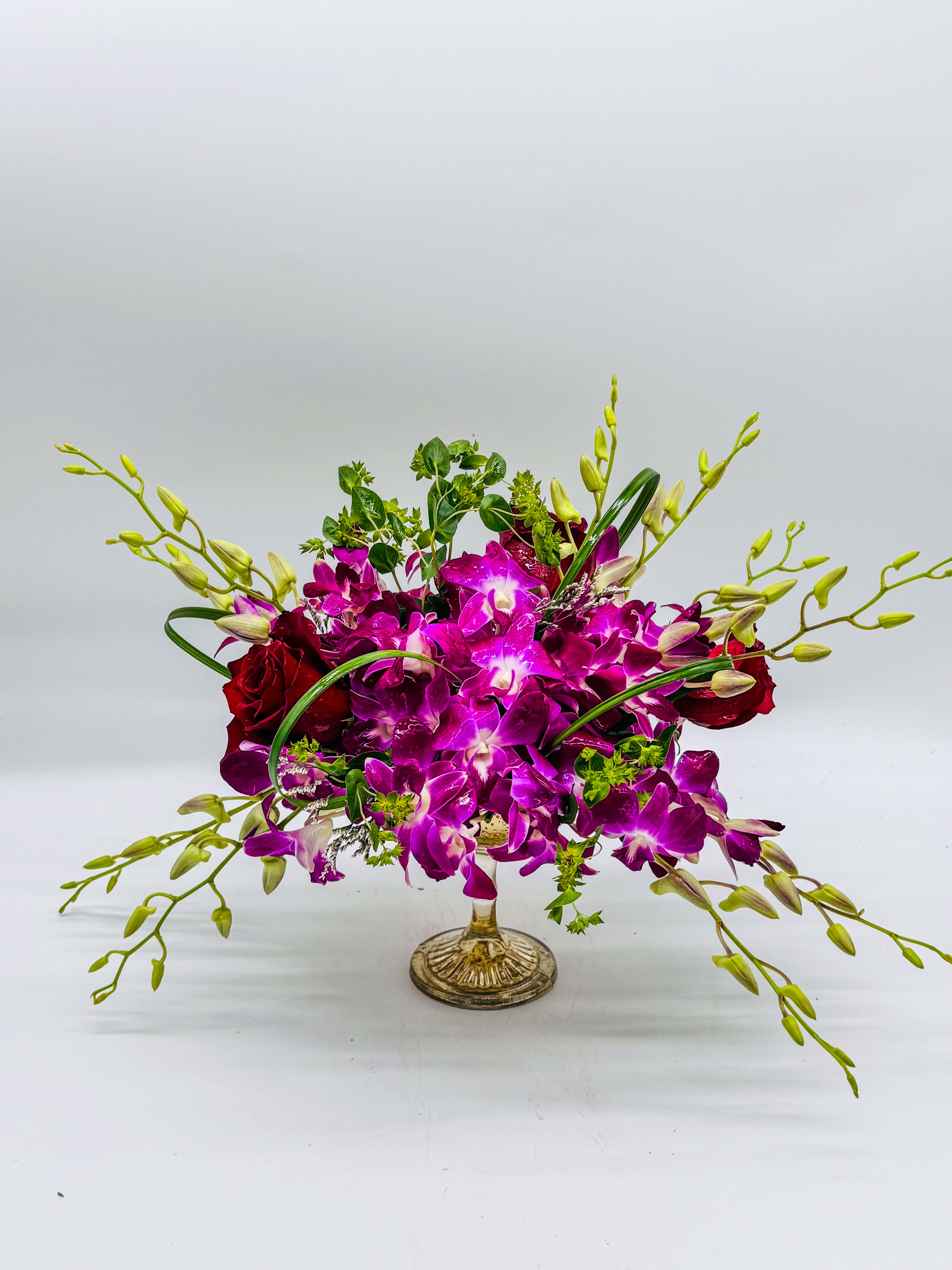 Violet Royalty - This is a stunning, low-profile centerpiece—think elegant dinner table or special-event vibe. A lush mix of deep purple dendrobium orchids (those big, velvety blooms) sit front and center, Red roses peek through like little jewels, while lime-green buds and curly stems add that fresh, wild edge. It’s all perched on a shiny gold compote dish, with wispy greens and tiny white fillers drifting out—like it’s floating. Super romantic, bold colors, zero clutter. Perfect for a wedding, anniversary, or just because.perfect for anniversaries, birthdays, F, get well, graduations or just because thank you , I'm sorry , Mother's Day,Valentine's Day