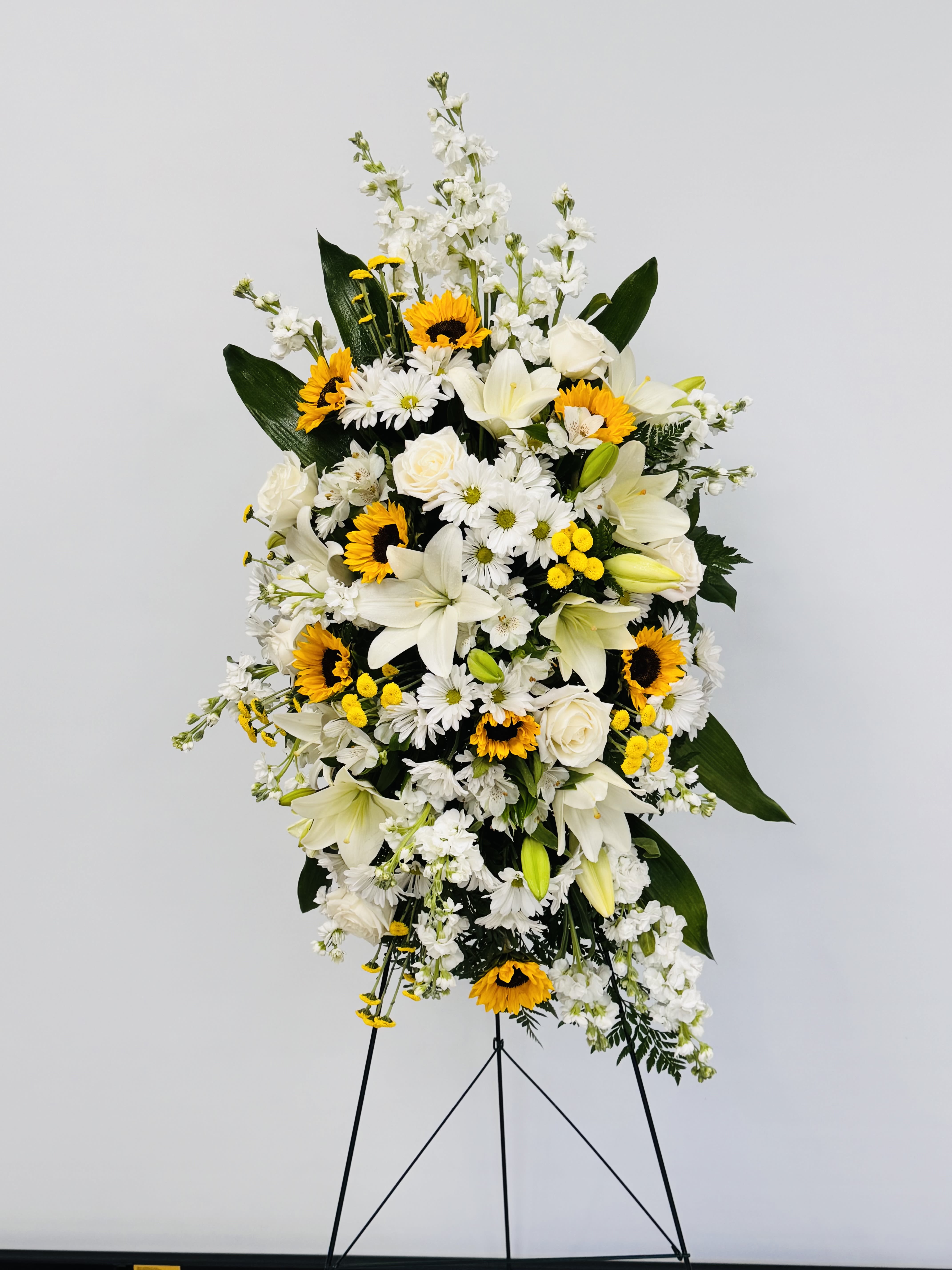 Sunlit Tribute Spray  - A radiant, reverent standing spray that balances consolation with bright, hopeful accents - perfect as a sympathy arrangement or memorial tribute. The tall arrangement reads formal and ceremonial on its metal easel stand, making it a dignified choice when you want the person you're honoring to feel remembered and celebrated. White lilies and roses deliver a peaceful, respectful presence while the sunflowers and yellow daisies add warm, uplifting highlights that suggest light and remembrance.  This spray features layered vertical lines of blooms: white lilies and roses woven with white daisies and alstroemeria, punctuated by clusters of vivid yellow sunflowers and small yellow accents. Broad, dark foliage gives depth and structure, framing the flowers so they read clearly from a distance during a service. The metal easel provides sturdy support and a classic presentation suited to funeral homes, memorial services, or graveside gatherings.  Made to convey sympathy with a comforting brightness, this arrangement is intended for honoring a life and offering solace to family and friends. The tall scale and easel mean it's best for standing-display needs; please tell us the service location and date when ordering so we can ensure timely delivery and proper setup.funeral 