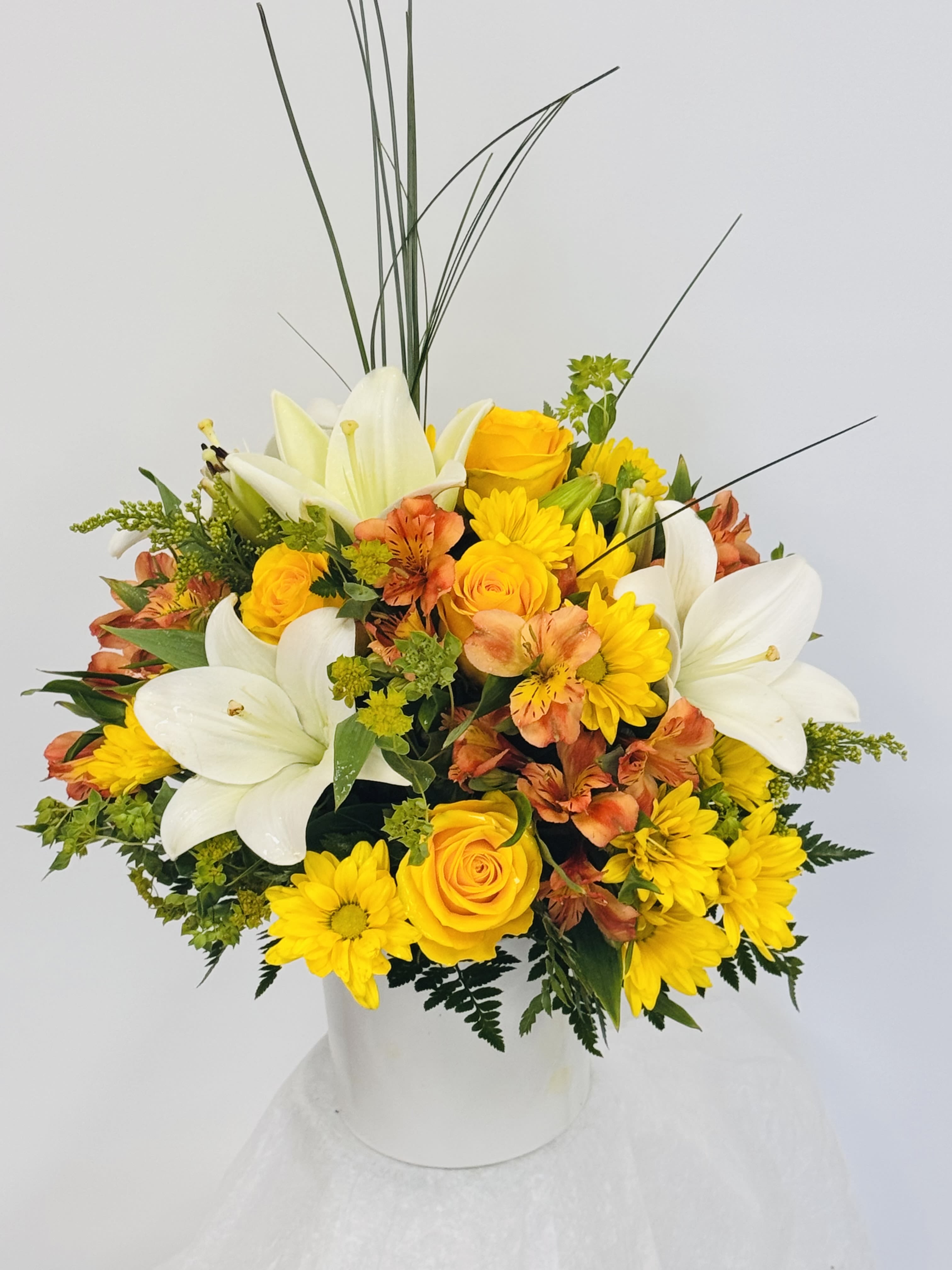 Sunshine Blaze. - Sunshine Blaze greets them like a burst of daylight-perfect as a cheerful birthday bouquet or an uplifting everyday arrangement to say congratulations or "thinking of you." The composition pairs large white lilies with bright yellow roses and sunny yellow chrysanthemums, the orange alstroemeria adding lively contrast so the recipient will instantly feel noticed and celebrated.  This rounded, full design sits in a clean white ceramic vase for a modern, versatile presentation. Tall, grass-like stems rise above the blooms to give height and graceful movement; overall the palette of yellow, orange, white, and fresh green foliage creates warmth and energy without being fussy. Ideal for a desk, dining table, or entryway, it brightens a room with bold color and soft lily fragrance.  Choose this arrangement when you want someone to feel genuinely cheered-whether for a birthday, a promotion, a get-well sentiment, or simply because today feels like a day to celebrate them. The compact rounded shape makes it easy to place, and the sturdy ceramic vase means it arrives ready to display.