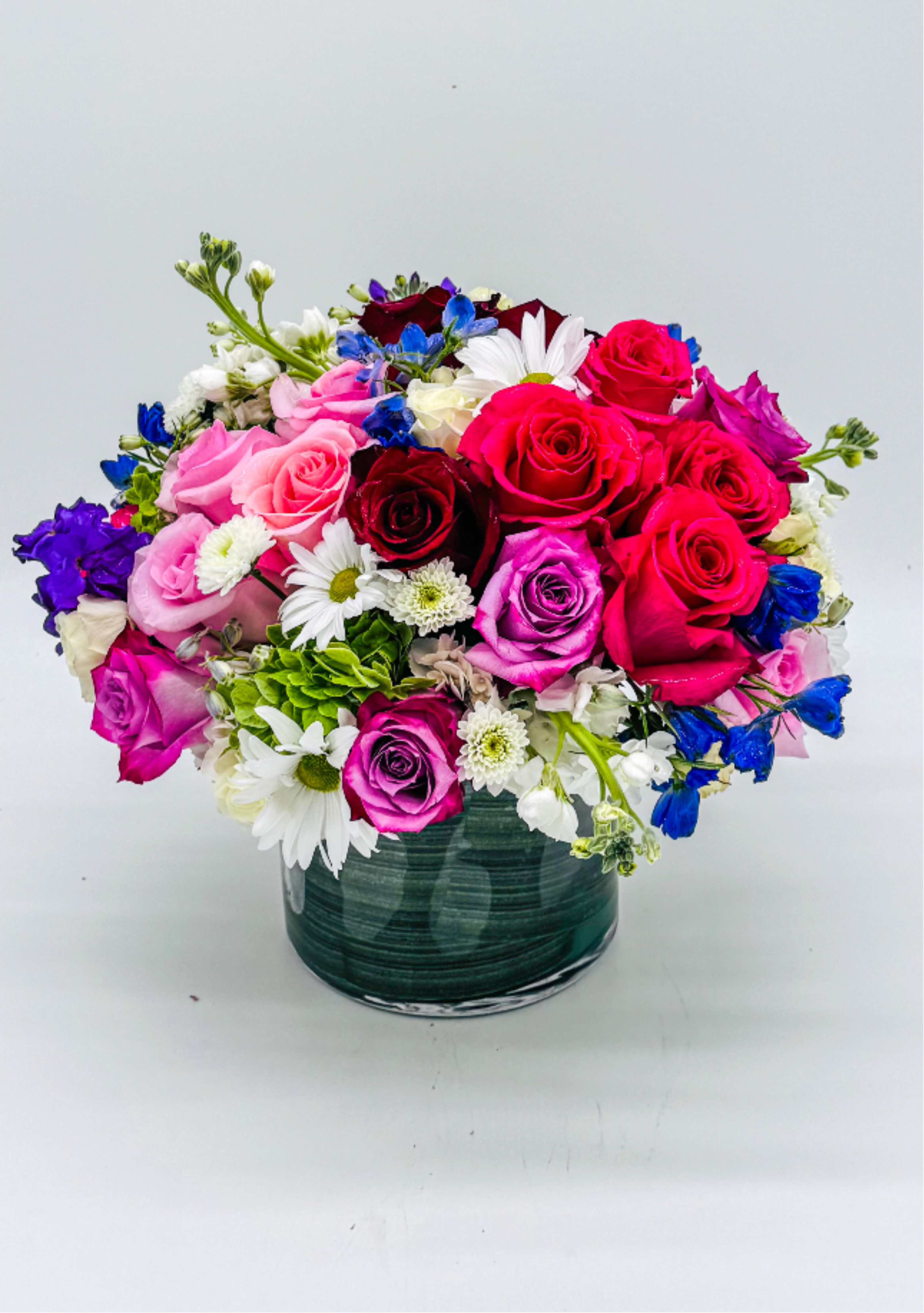 Floral  - A vibrant, round floral arrangement featuring a mix of pink, red, and purple roses, accented with white daisies and small blue blooms. The blooms are artfully arranged in a dark green, glossy container, creating a lush, romantic centerpiece with a fresh, cheerful aesthetic.perfect for anniversaries, birthdays, Father's Day, get well, graduations or just because thank you , I'm sorry , Mother's Day,Valentine's Day