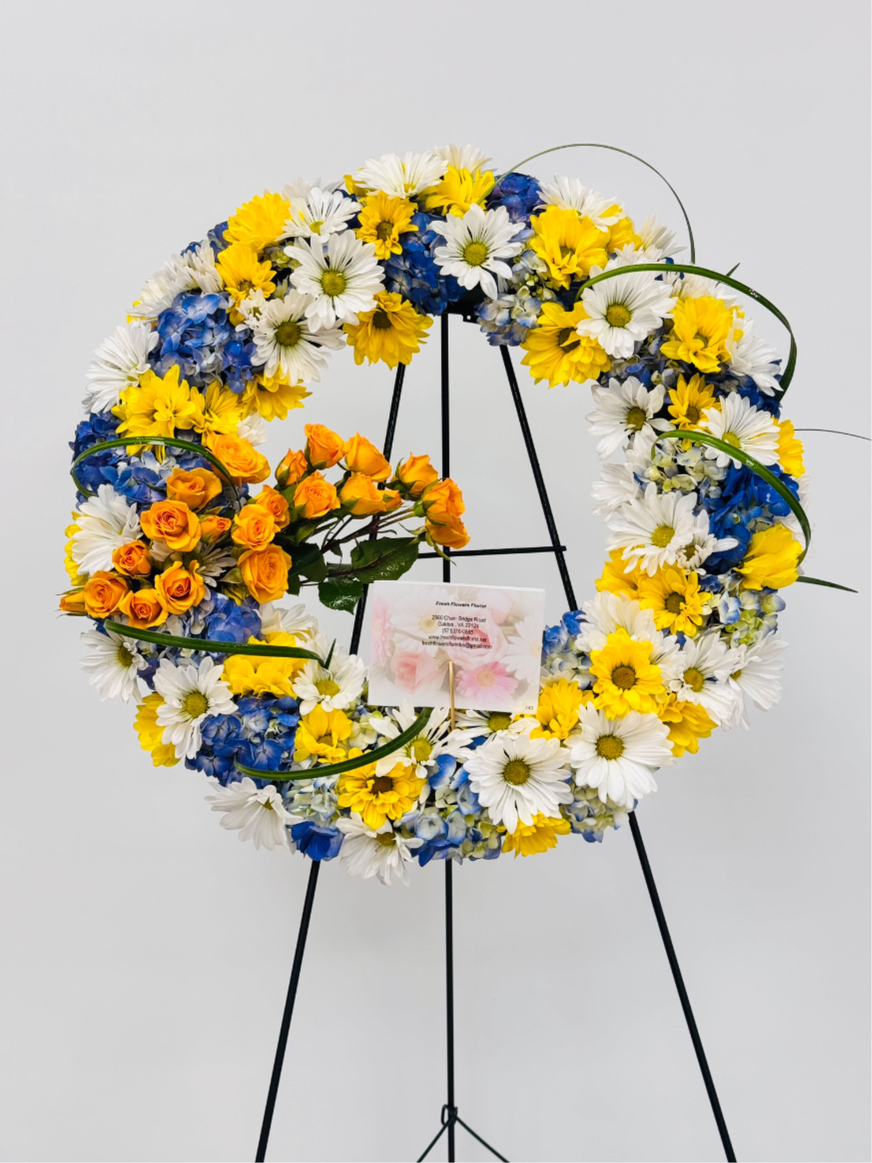 Heavenly Tribute - A bright circular wreath that reads as both celebratory and respectfully elegant, this flower arrangement is designed for memorial services or congratulatory displays. The wreath form and metal easel stand give it a formal presence ideal for a funeral tribute, remembrance, or ceremony where you want the person you're honoring to feel celebrated and remembered. The mix of yellow, white, blue, and orange flowers creates an uplifting palette that catches the eye without feeling gaudy.  Carefully wired on a black wreath stand, the ring is densely filled with white and yellow daisy-like blooms that form a sunny, textured backdrop. Cool blue hydrangea clusters are placed throughout the circle to add visual depth and contrast, while a focused spray of small orange roses on the lower left provides a warm, asymmetrical focal point. The arrangement arrives ready to display on the included metal easel and comes with a card for a personalized message.  Appropriate for a wide range of occasions-from a heartfelt sympathy tribute to a proud celebration of life or achievement-this wreath communicates warmth and respect for the recipient. Size and presentation are tailored for public display; please indicate any special wording for the card when ordering so we can prepare it for delivery and placement.funeral and sympathy 