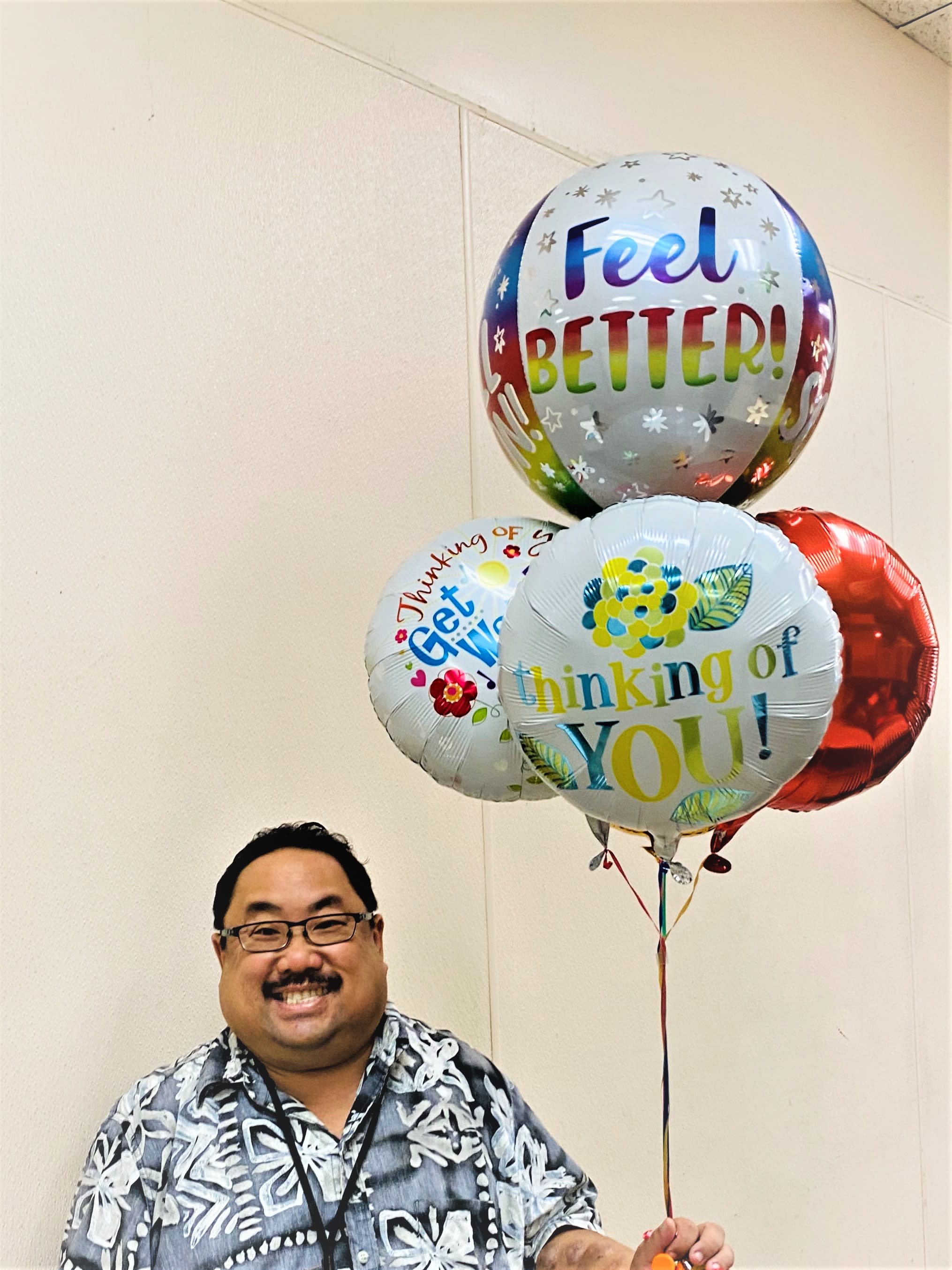 Smiling man holding three colorful get well message balloons in front of a beige wall