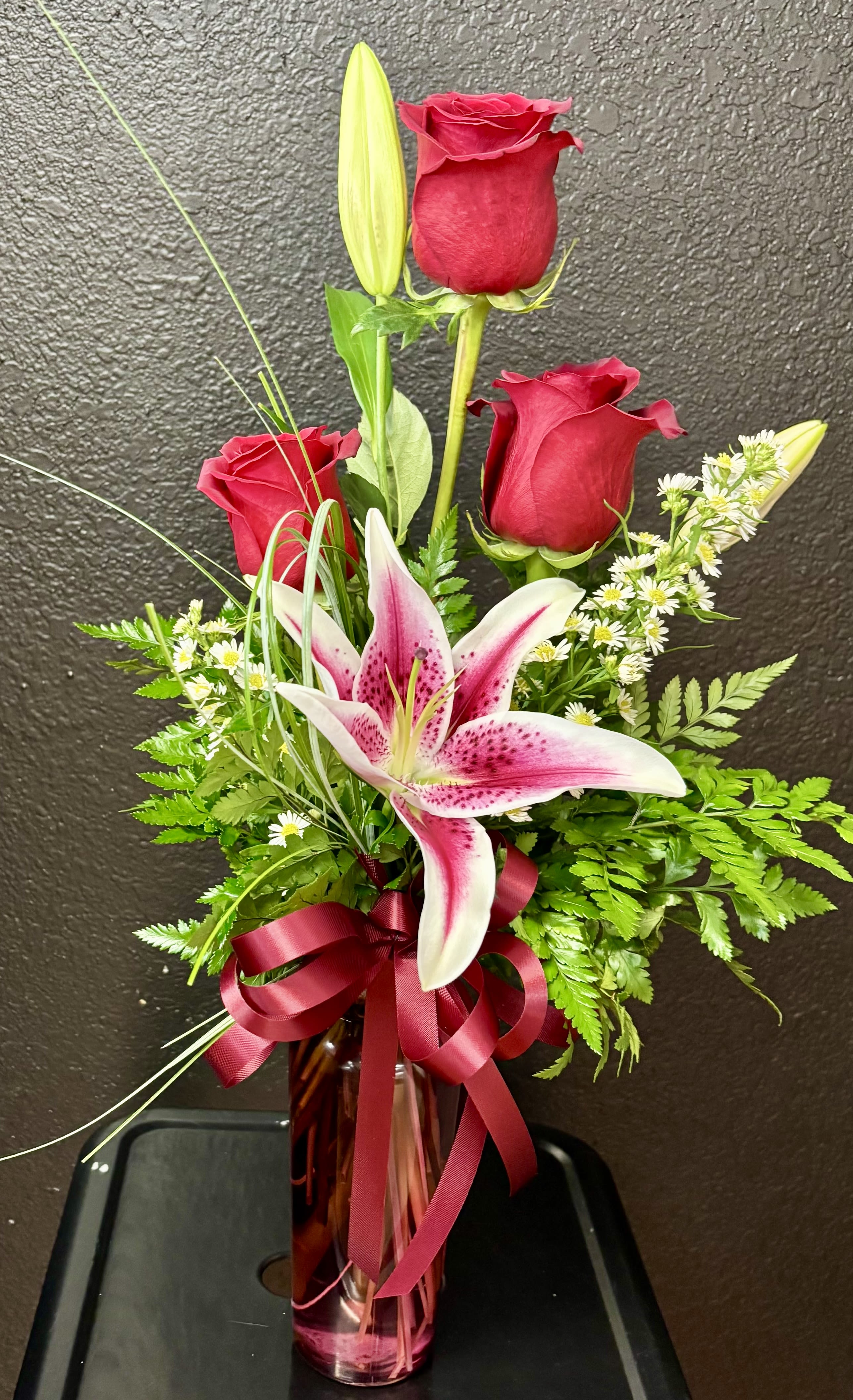 Tall arrangement of red roses and a pink stargazer-style lily in a red glass vase with ribbon