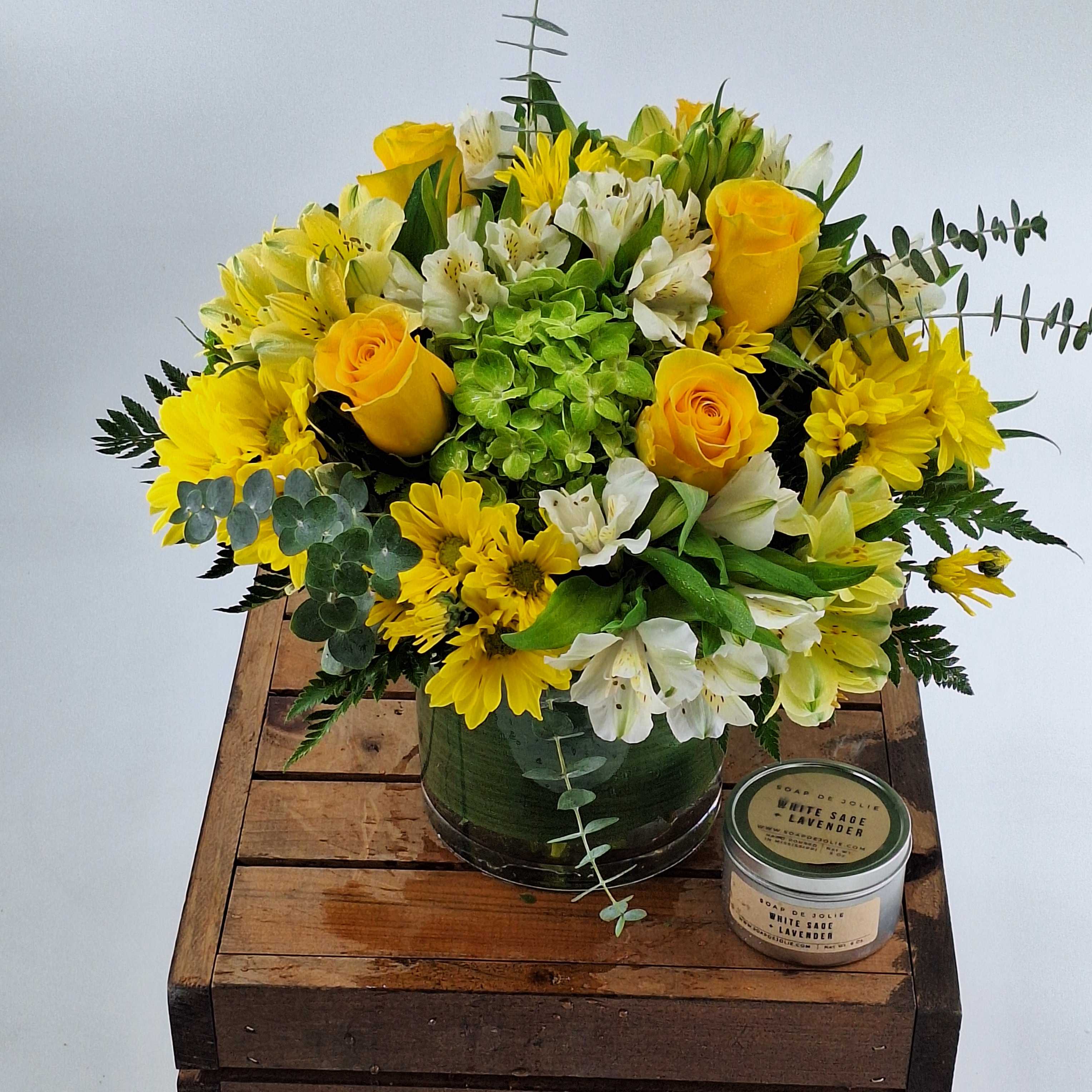 Yellow roses and white flowers in a green vase with a candle beside it.
