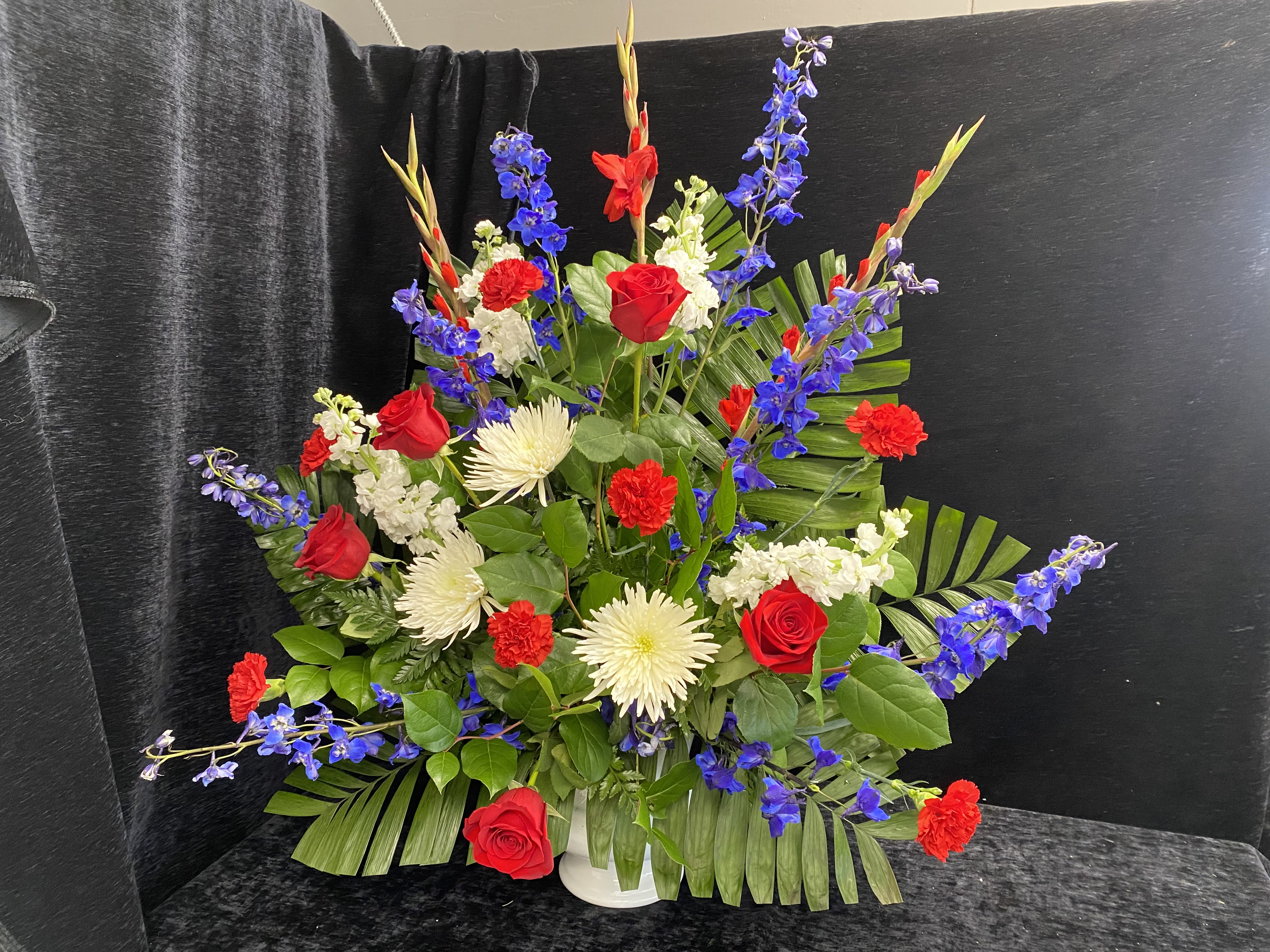 Patriot Funeral Spray - A bold upright spray of red roses, red gladiolus, white spider mums, vivid blue delphinium, red carnations, white stock and lush greenery makes a powerful sympathy and funeral tribute that reads patriotic without feeling gimmicky. The red, white and blue palette gives the arrangement a formal, respectful presence-ideal for Memorial Day, a funeral service, or to honor someone who served-while still working as a confident sympathy gift that says "we remember" or "you are in our thoughts." The sturdy white vase and lush foliage help keep stems supported; water upon delivery and refresh as needed to maintain peak appearance through the days following delivery.