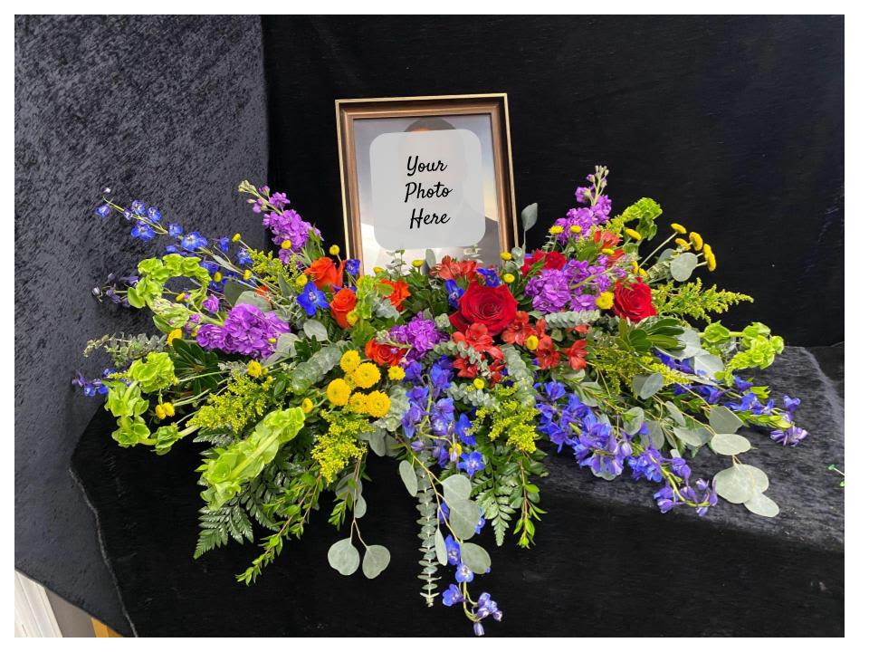 Celebration of Life Photo Spray - A colorful, commemorative photo spray designed for remembrance or a funeral tribute. This celebration-of-life arrangement centers around your framed photo, surrounded by a low, horizontal spread of mixed-color blooms. The design suits a memorial service or a special commemorative display, offering a bright, hopeful alternative that honors a life with warmth and dignity.  This arrangement can be displayed on top of a casket or on a table. Photo and frame provided by the customer.