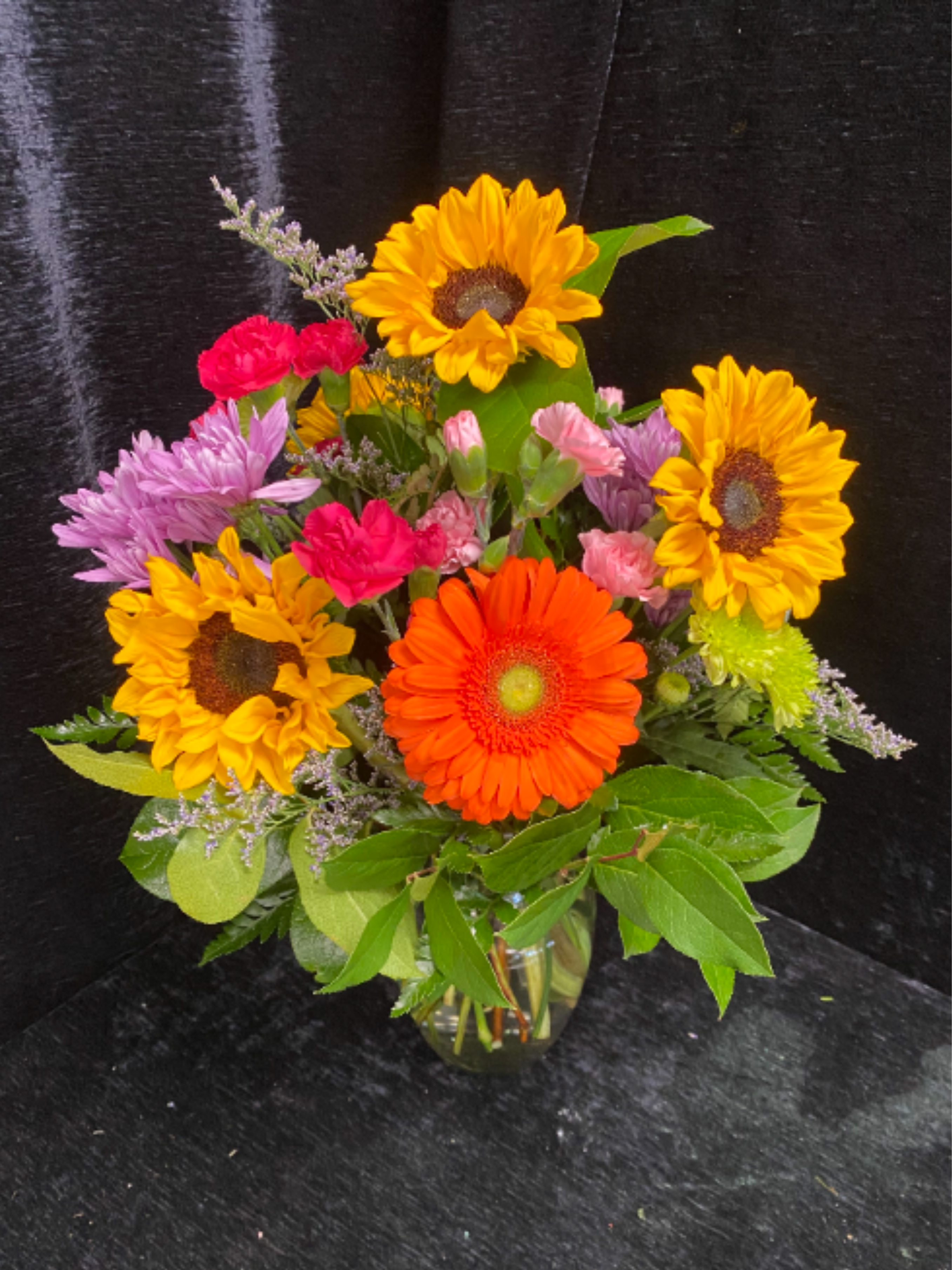 Bright mixed bouquet with sunflowers, orange gerbera, carnations, and mums in a clear glass vase