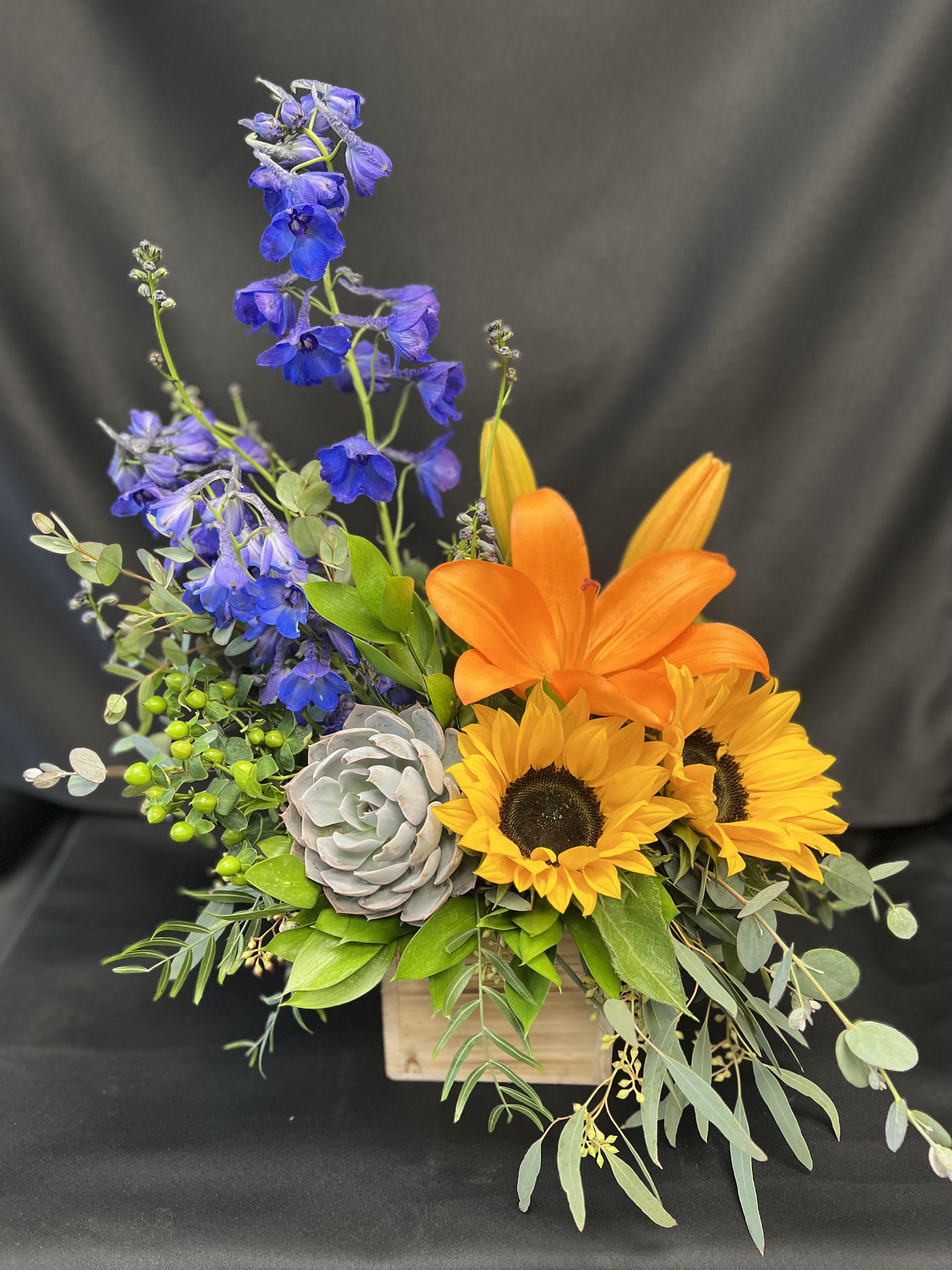 A Ray of Sunshine - Summery, colorful - like a ray of sunshine. The wooden box and succulent adds a bit of a relaxed feel to this beautiful bouquet. Colorful lilies, sunflowers and deep blue delphinium in a perfect blend.