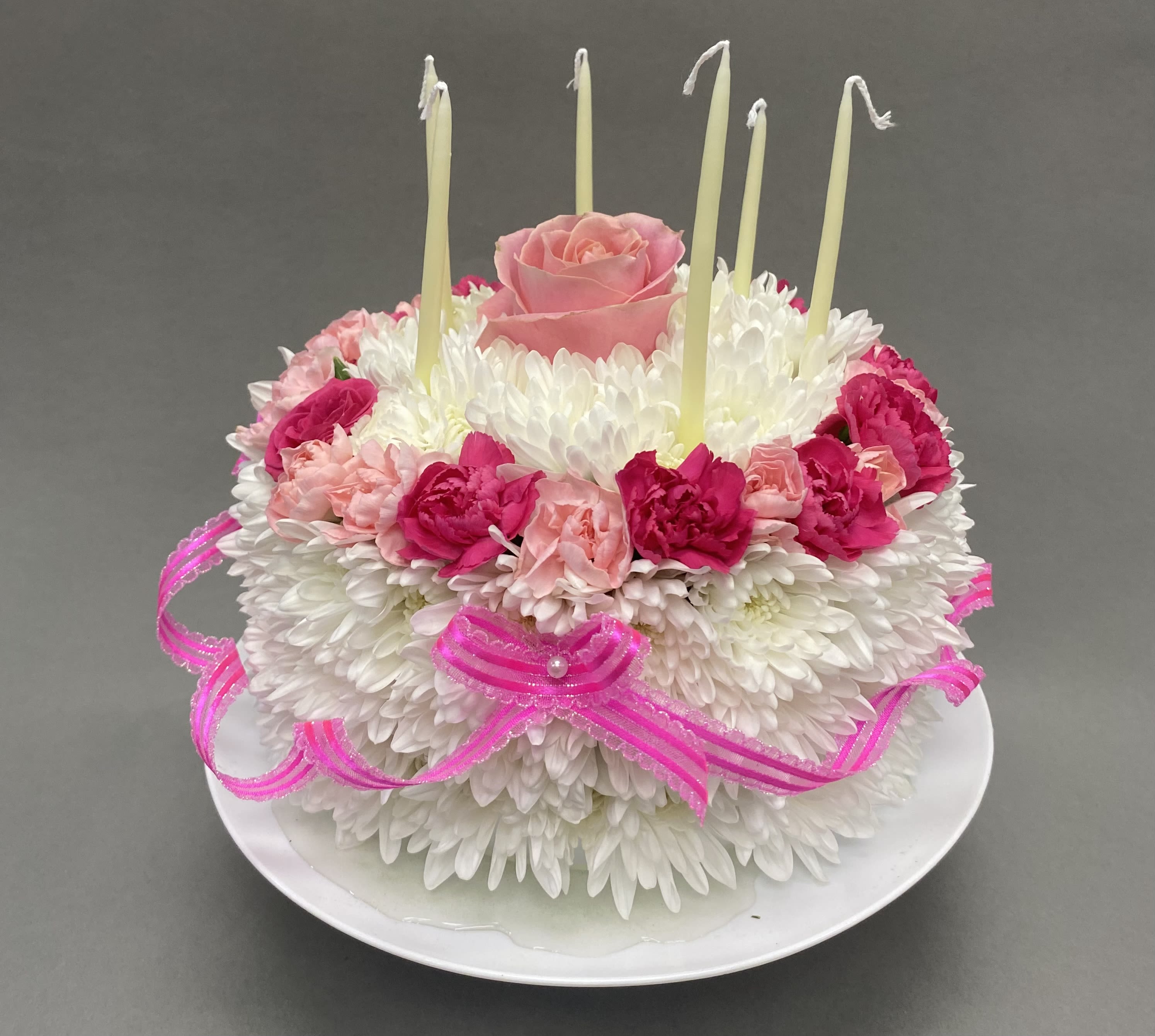 Round floral cake of white mums with pink roses, carnations, and tall candles on a plate