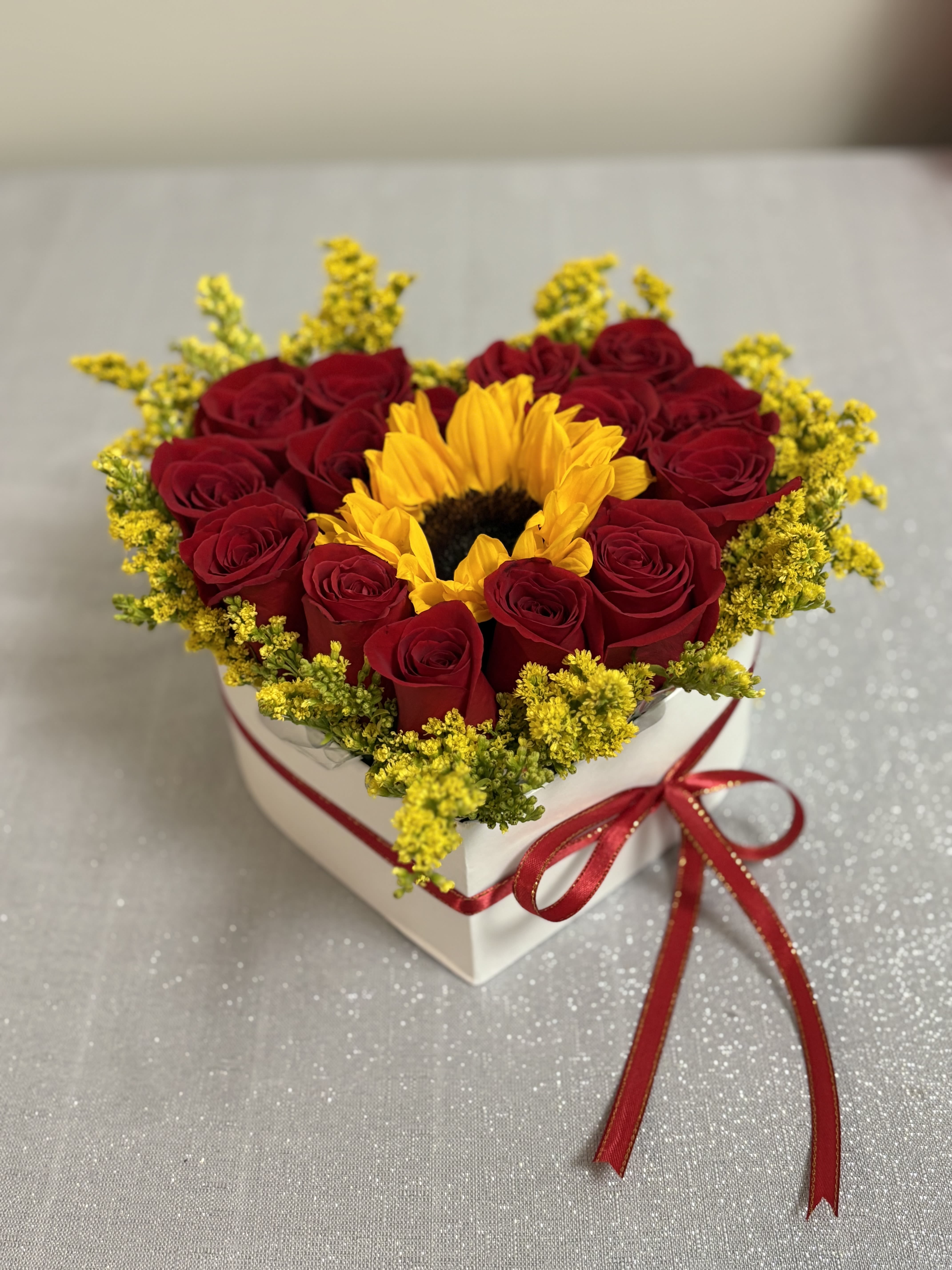 Heart-shaped box of red roses with a sunflower center and red ribbon