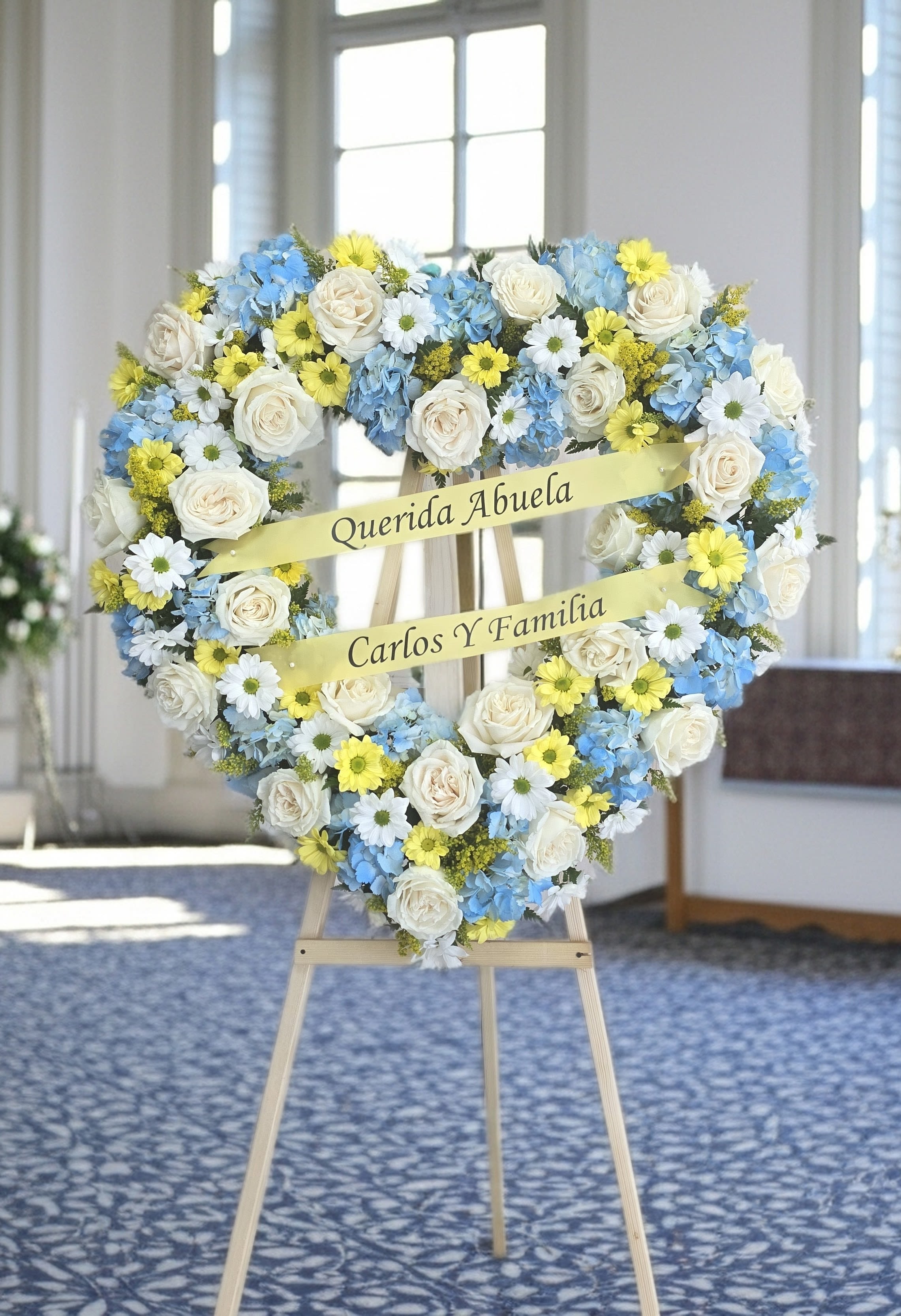 Soft Serene Heart Spray - Banner not included* A large heart-shaped standing spray in soft whites, sky blues, and gentle yellows-this sympathy heart spray makes a tender memorial for a funeral or celebration of life. it's the kind of tribute that lets the person you're honoring feel remembered and cherished.  Hand-arranged on a sturdy wooden easel, the spray blends cream-white roses with clusters of blue hydrangea and small white-and-yellow daisy-like blooms for a mixed-color, comforting palette. The texture moves from the full, cloud-like hydrangeas to the classic rose petals and the delicate daisy accents; two pale yellow ribbons across the center add a gentle focal point. The overall size is large and designed to stand prominently at a service, offering clear visibility and a formal, respectful presence.  Made to convey sympathy and remembrance, this heart spray is suited for graveside services, viewings, or as a statement piece at a memorial gathering-the recipient's family will feel sincerely honored. 