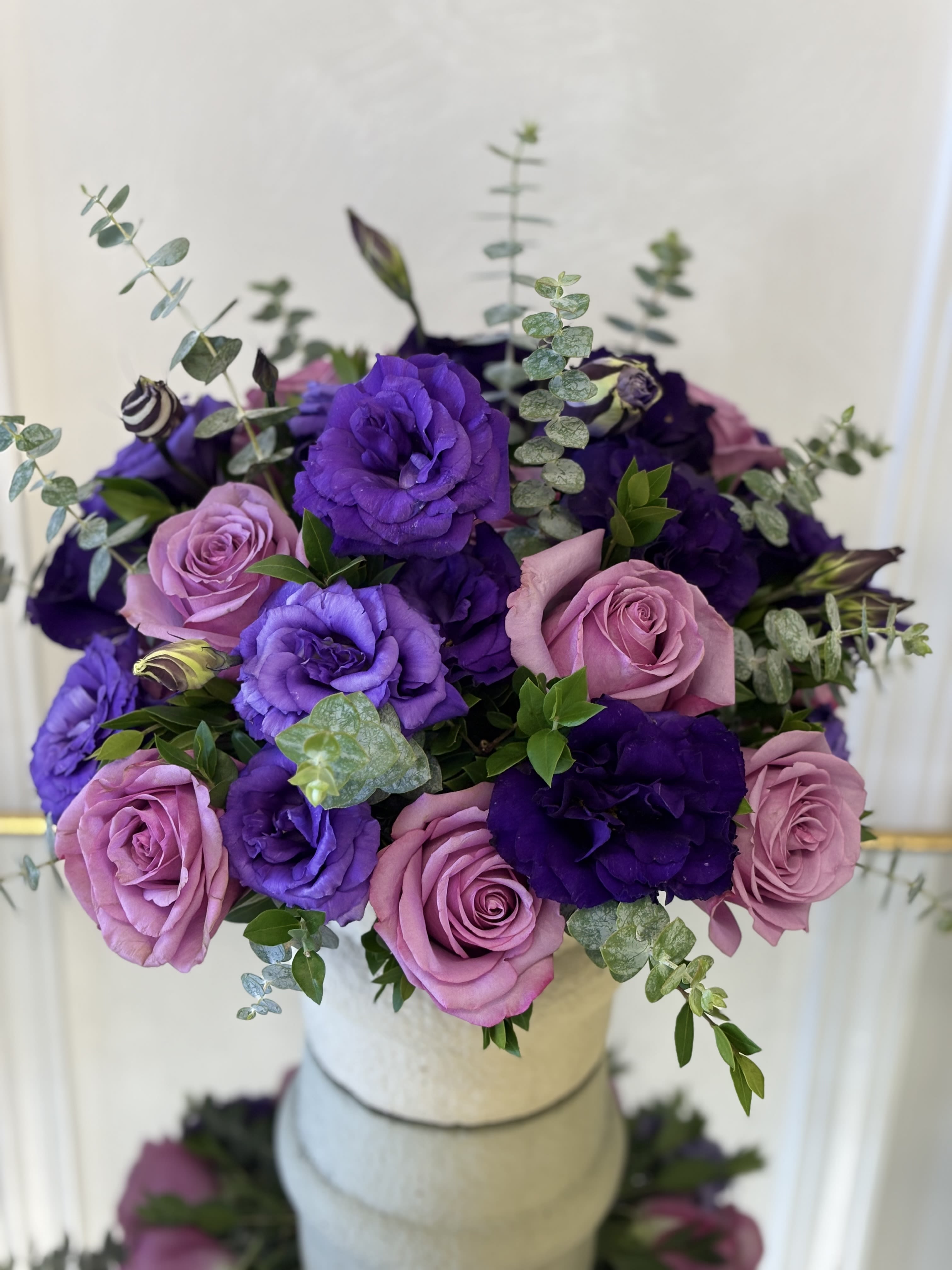 Purple Majesty By VLM Flowers - A luxurious arrangement featuring rich purple lisianthus, deep violet blooms, and soft lavender roses accented with fresh eucalyptus and lush greenery. Designed with a romantic blend of elegance and charm, this timeless bouquet brings a touch of royal beauty and sophistication to any space.