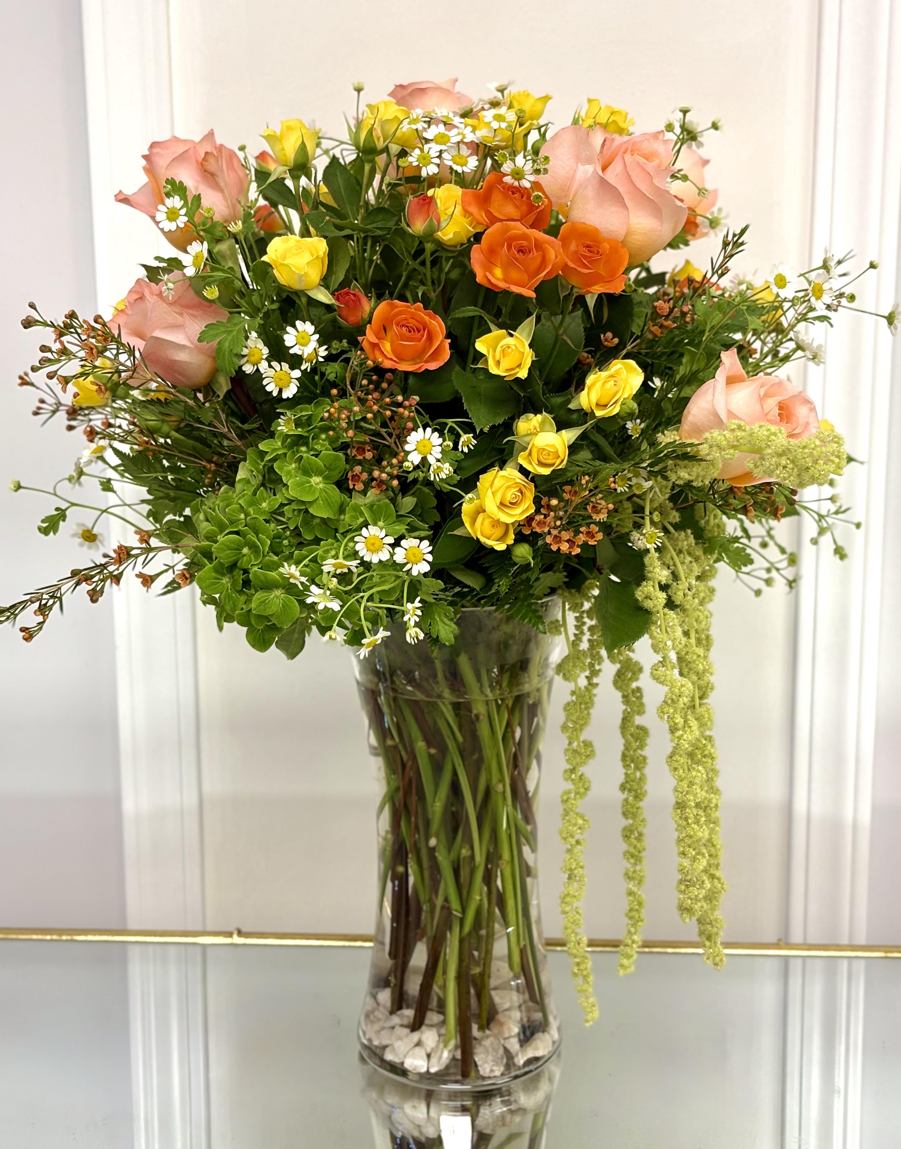 Golden Garden Delight By VLM Flowers - Bring the beauty of a blooming garden indoors with this luxurious floral arrangement. Featuring elegant blush roses, vibrant orange and yellow spray roses, lush green hydrangea, delicate chamomile flowers, and cascading seasonal greenery, this handcrafted design is filled with texture, movement, and natural charm. Arranged in a clear glass vase, Golden Garden Delight is perfect for birthdays, anniversaries, congratulations, housewarmings, or simply celebrating life's everyday moments with color, warmth, and elegance.