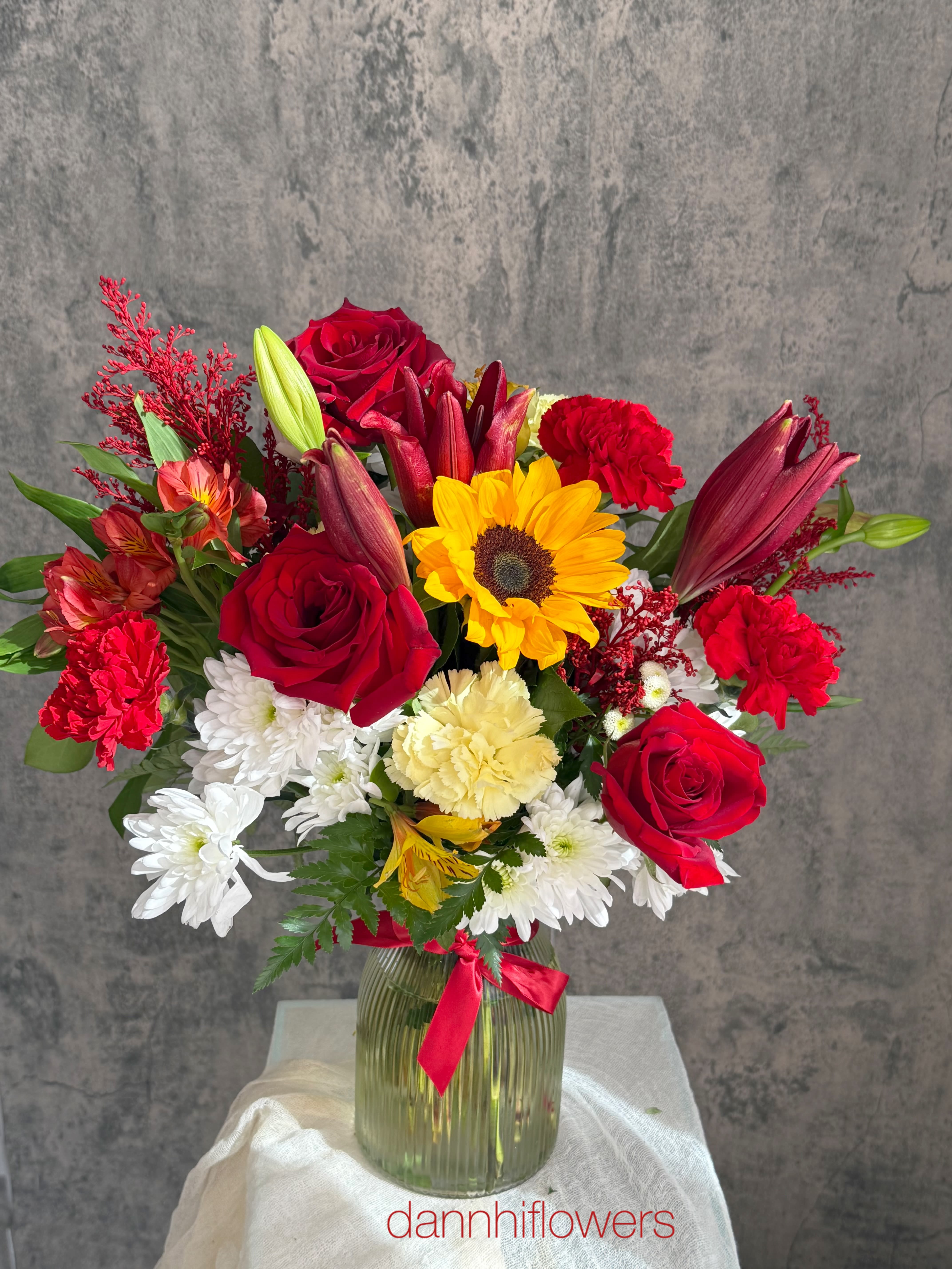 Mixed bouquet of red roses, lilies, sunflowers, and white daisies in a glass vase