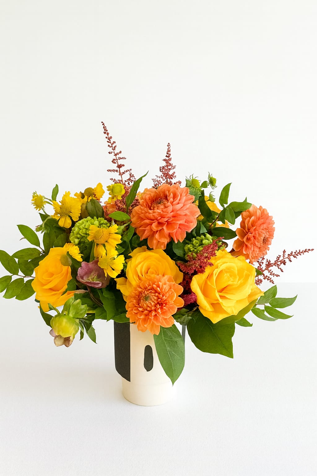 Golden Sunset - Bring the warmth of a late summer sunset into any space with this vibrant design. Golden yellow roses, cheerful yellow daisies, and bright orange dahlias come together in a joyful harmony, accented with fresh greenery and soft touches of burgundy astilbe. Designed in a modern black-and-white ceramic vase, this arrangement is both contemporary and timeless.  Perfect for brightening a home, celebrating a special occasion, or sending cheer, the Golden Sunset arrangement radiates happiness and warmth with every bloom.  Size: Approximately 8” W x 10” H Best for: Birthdays, celebrations, thank you gifts, or everyday joy