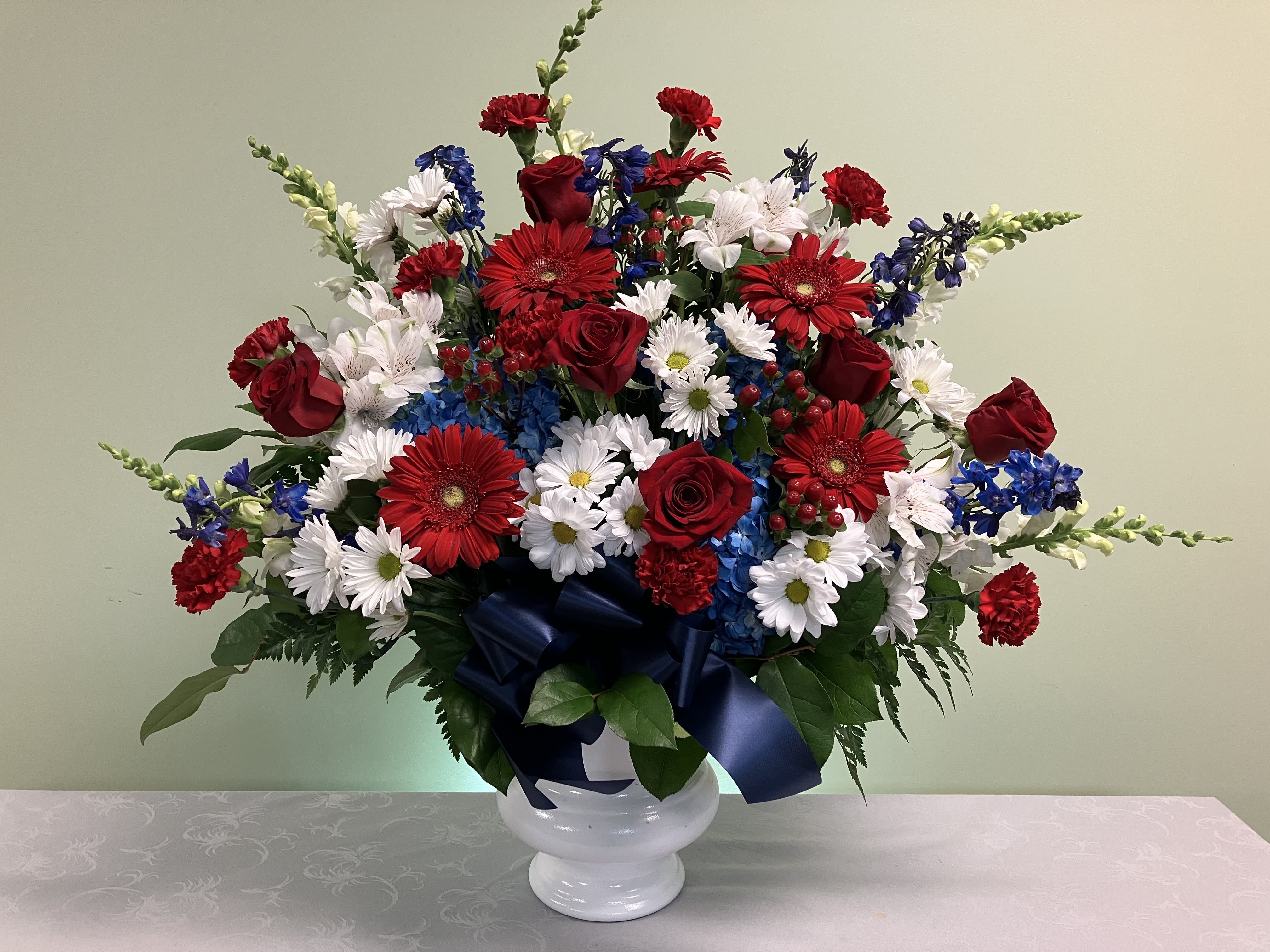 Patriotic red, white and blue flower arrangement in a white urn with a navy bow.