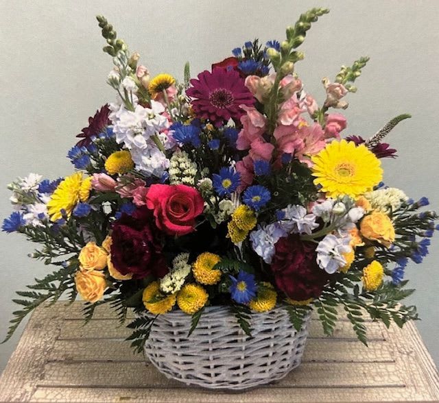 Mixed bouquet in a white wicker basket with bright yellow, pink, blue, and white flowers