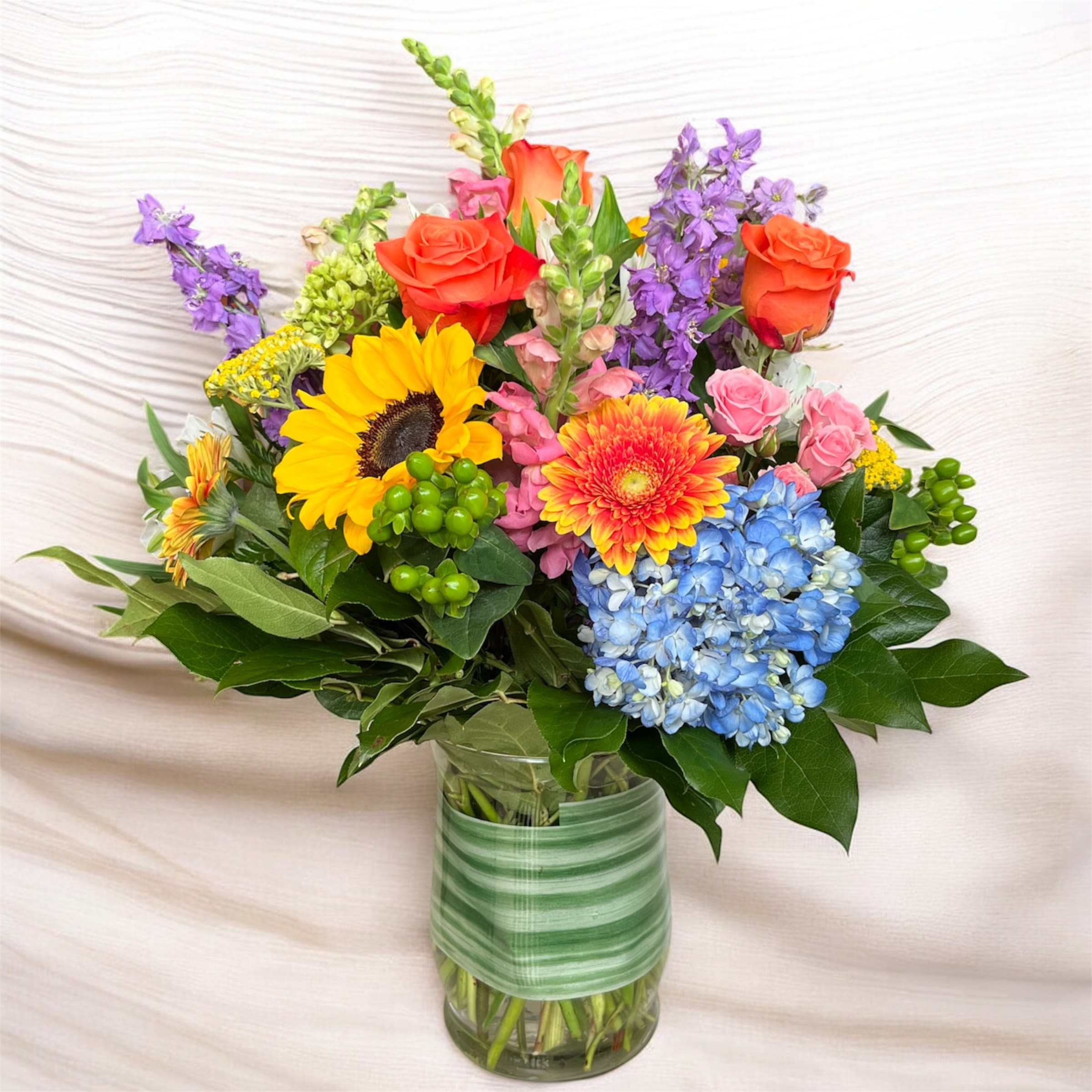ED18 Warm Horizons - Tall and Colorful this bright mix of floral is designed in a clear glass vase. Upgrading to a Deluxe or Premium arrangement will add more blooms to the design.
