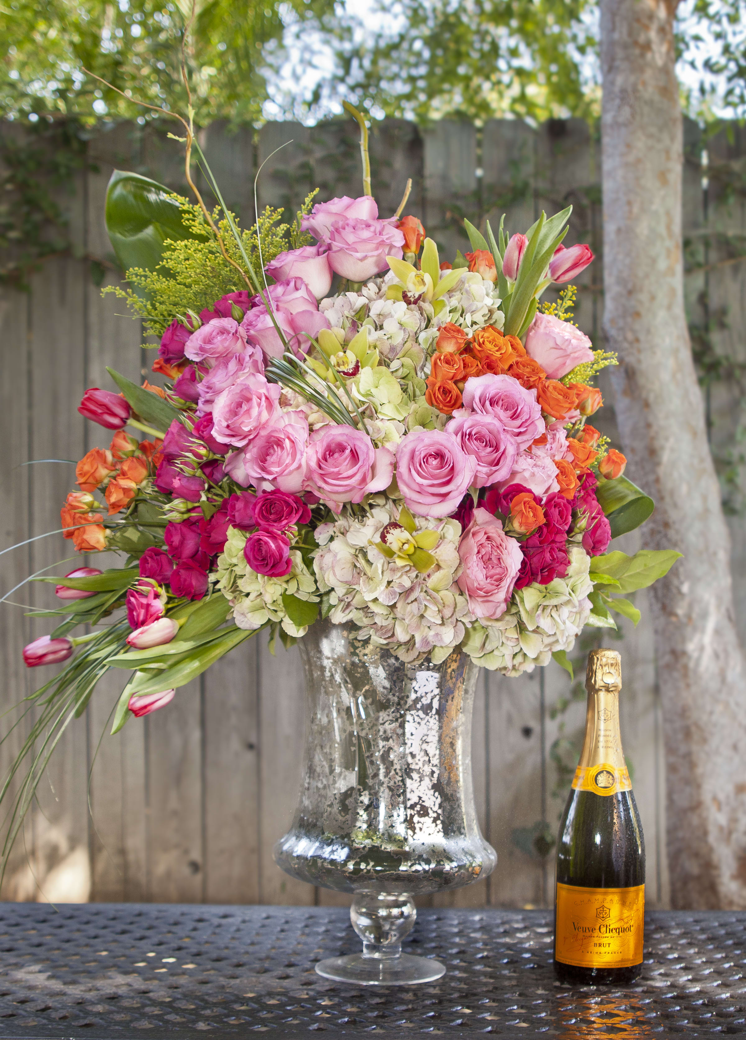 Large pink and orange floral arrangement in a glass vase beside a champagne bottle