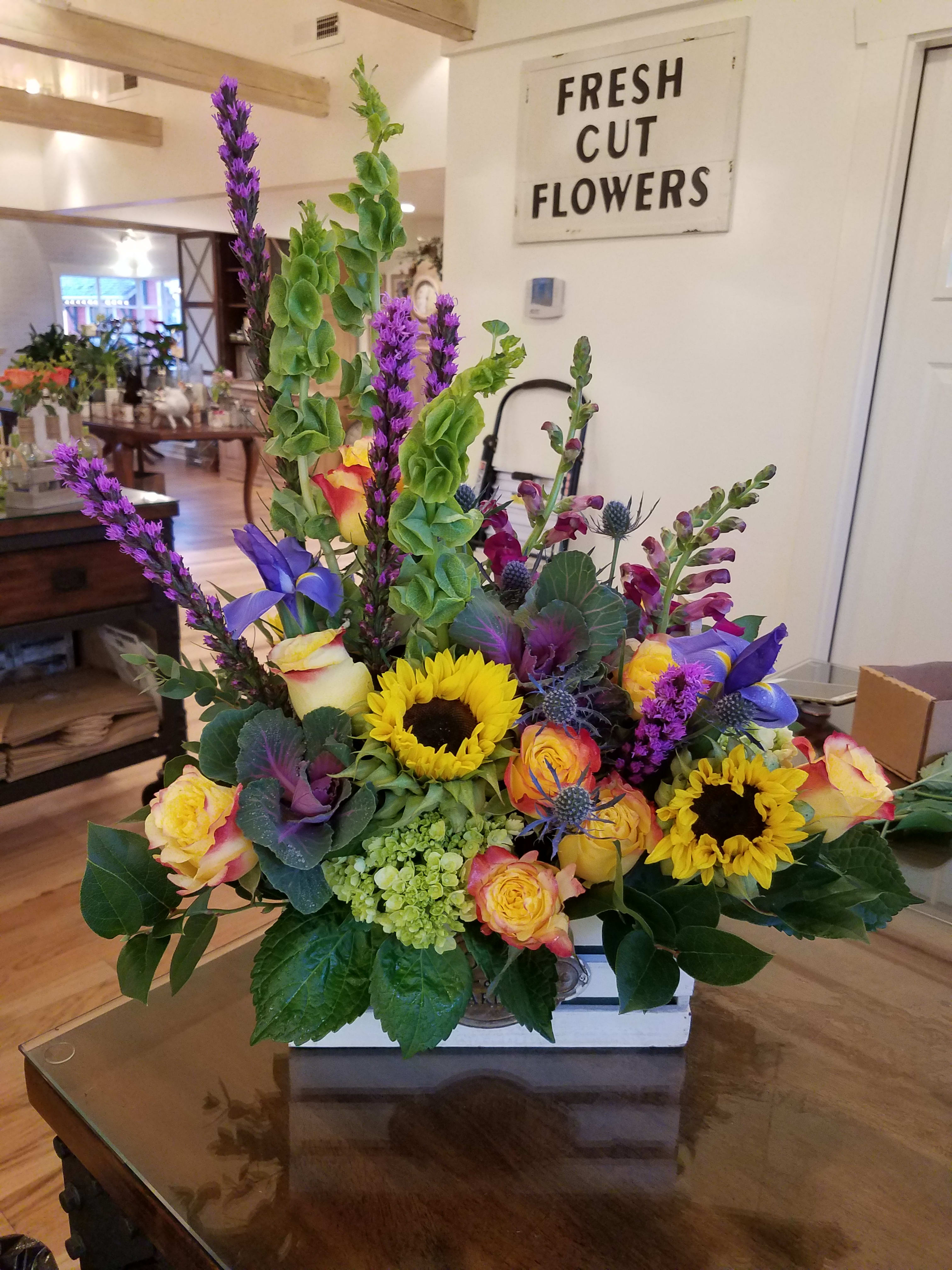 Bright mixed arrangement with sunflowers, roses, and purple flowers in a white wooden box.