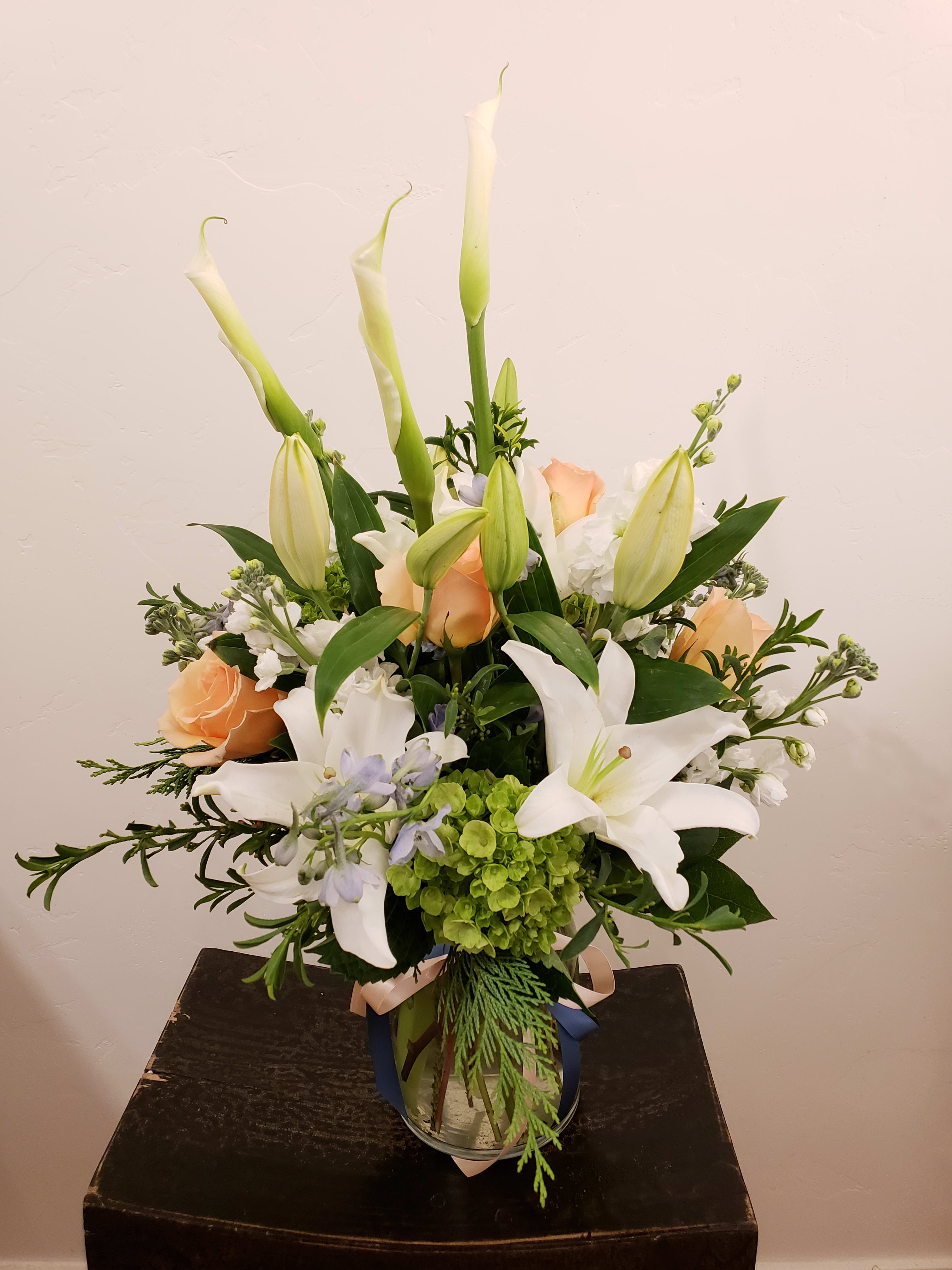 Tall arrangement of white lilies and peach roses with green hydrangea in a clear glass vase