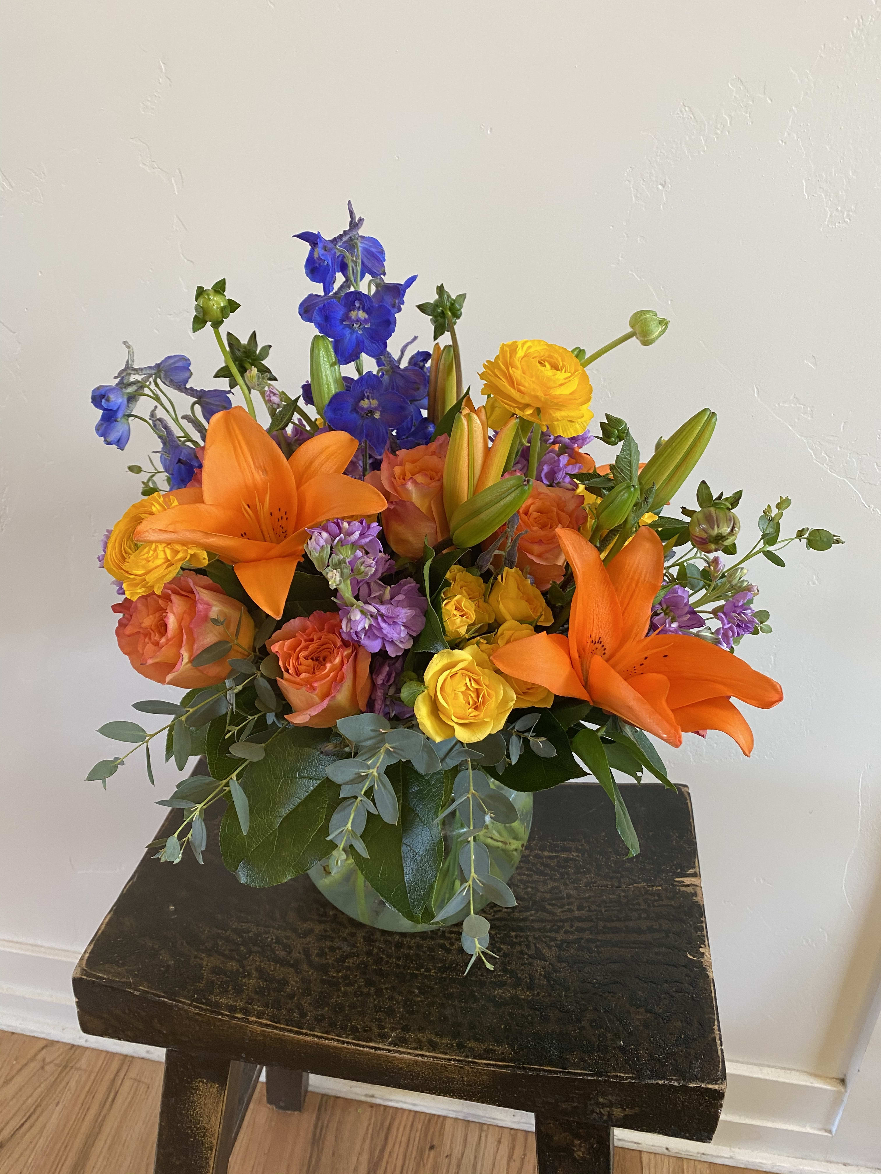 Bright mixed bouquet with orange lilies, roses, and blue flowers in a glass vase