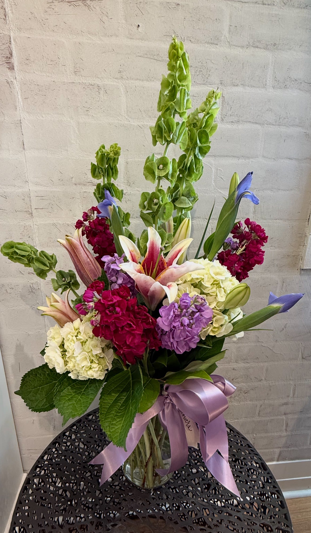 Tall mixed floral arrangement with lilies, hydrangeas, and Bells of Ireland in a glass vase with a lavender bow.