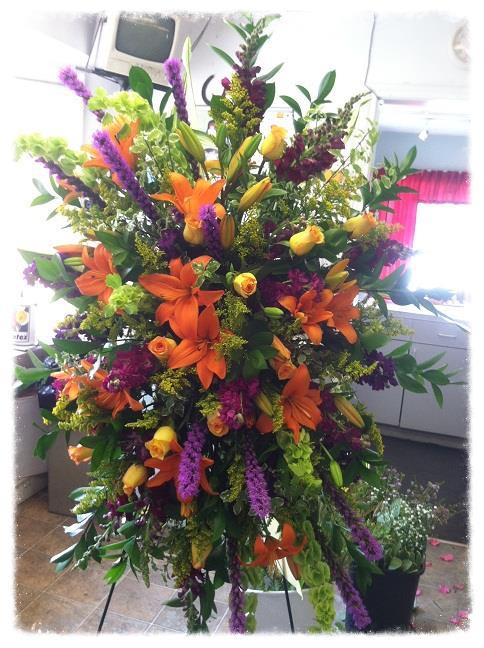 Tall standing spray of orange lilies, yellow roses, and purple flowers on a metal easel