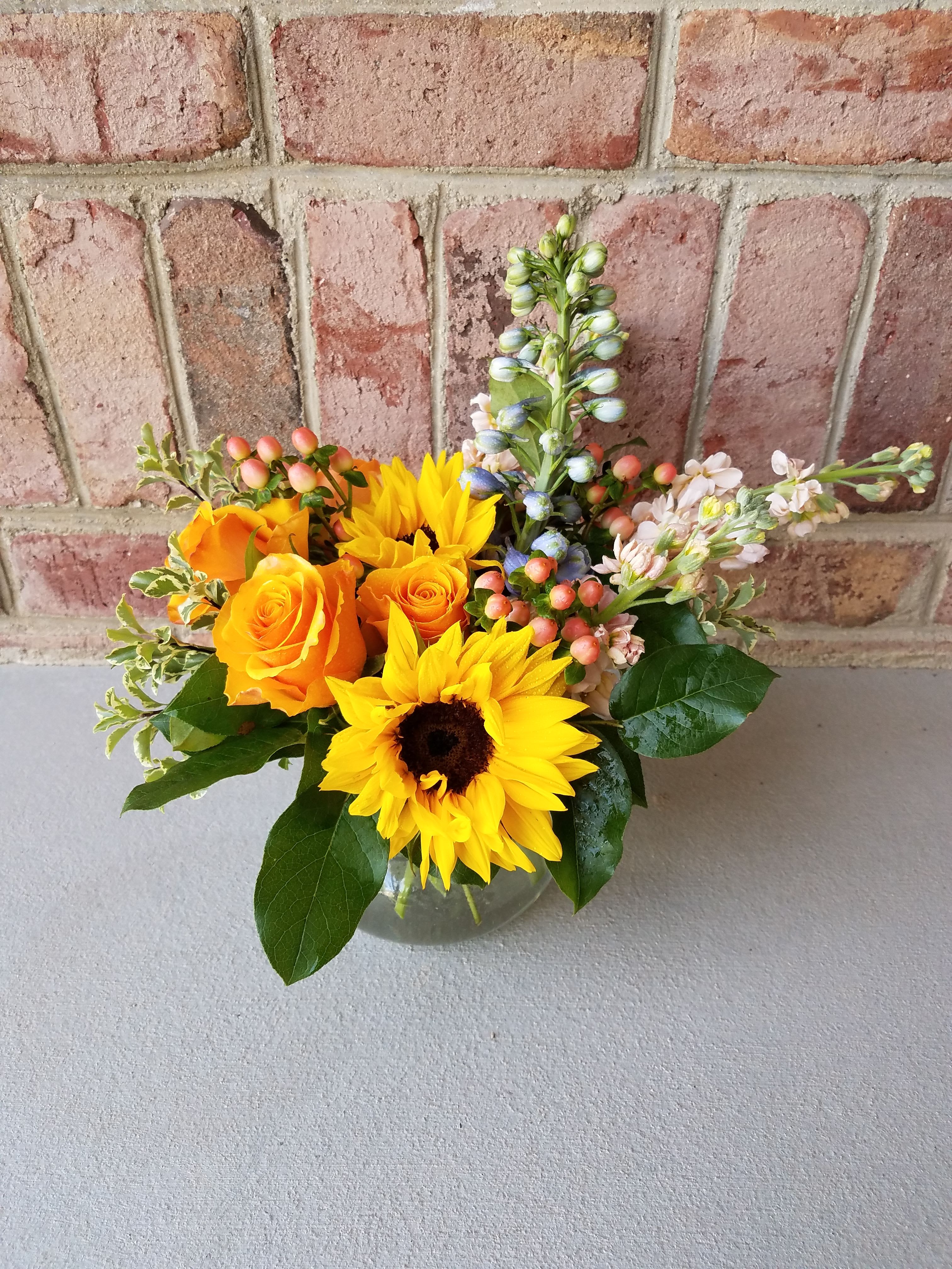 Low glass vase of yellow sunflowers, orange roses, and mixed accent blooms.