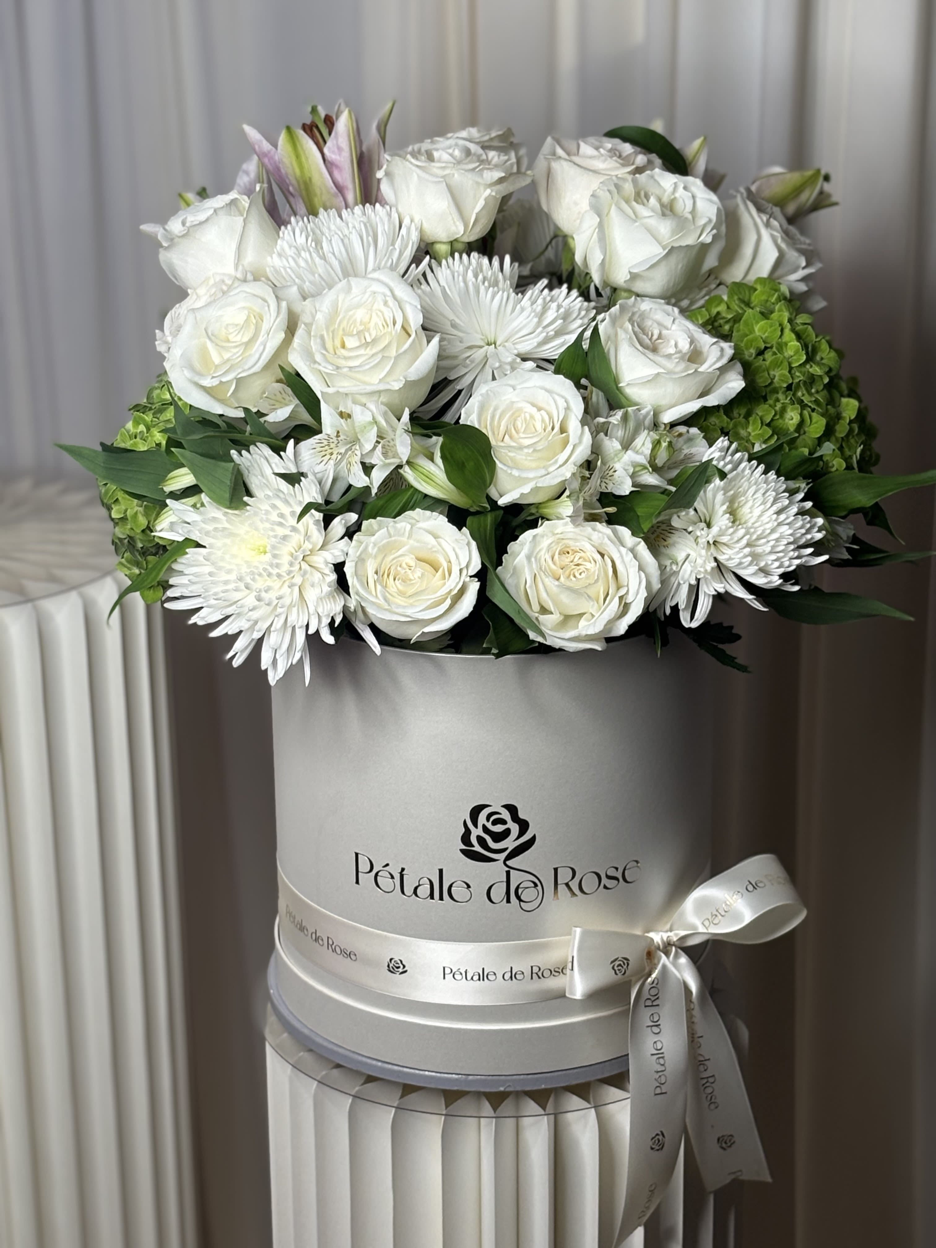 Royal White Box - A timeless arrangement designed to bring elegance, peace, and sophistication to any occasion. Featuring premium white roses, fragrant lilies, white spider mums, alstroemeria, and seasonal hydrangeas, this luxurious hat box is carefully handcrafted to create a refined and unforgettable floral statement.  Perfect for birthdays, anniversaries, sympathy, congratulations, housewarmings, or simply to brighten someone’s day with timeless beauty.  Flowers may include:  * Premium White Roses * White Lilies * White Spider Mums * White Alstroemeria * Hydrangeas * Premium Seasonal Greenery  To ensure the freshest and highest-quality flowers, some blooms or foliage may be substituted based on seasonal availability. Any substitutions will always maintain the overall style, color palette, size, and value of the arrangement while preserving the luxurious design you selected. 
