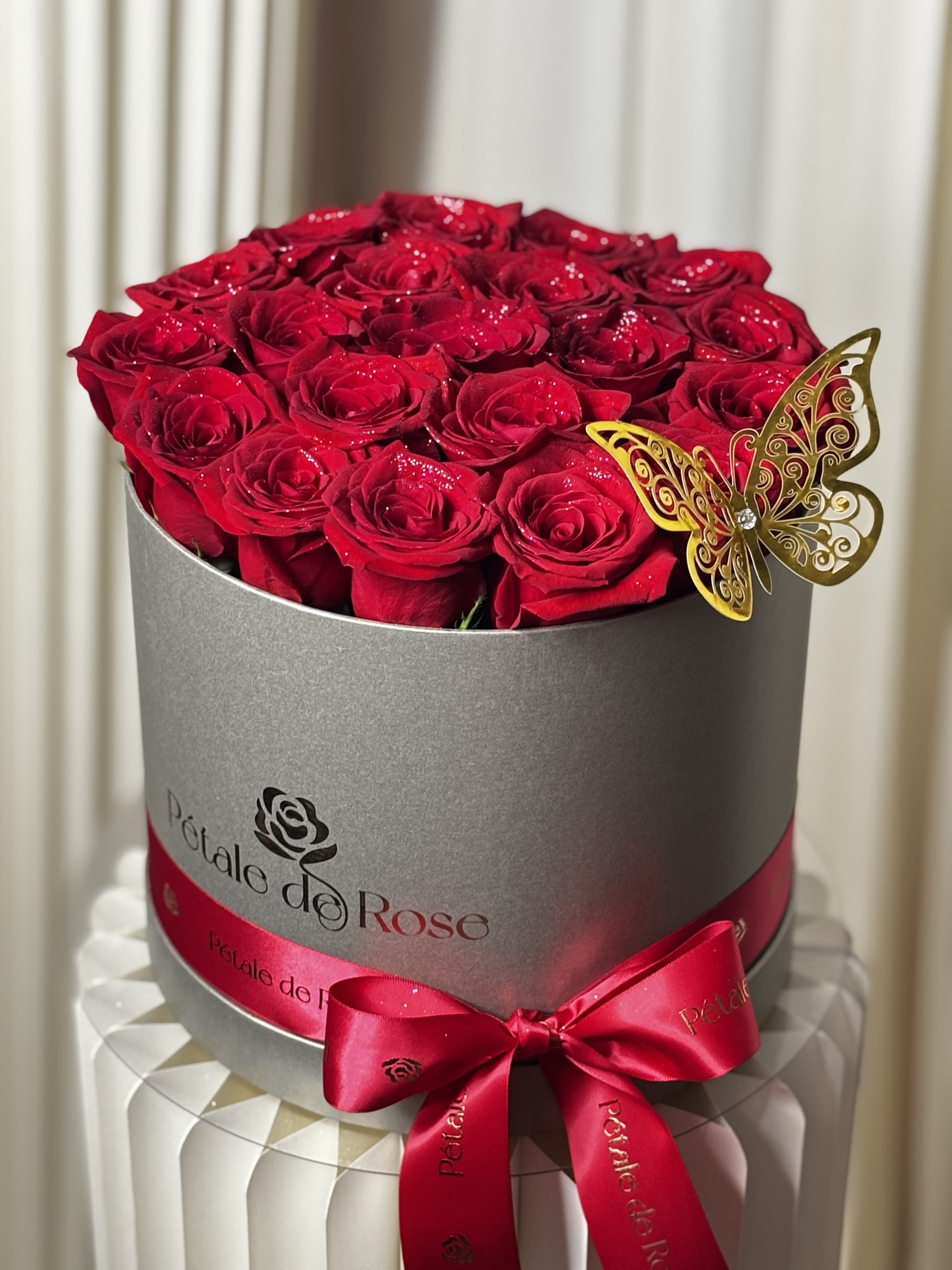 Ruby Royale Box  - A luxurious arrangement designed to make every moment unforgettable. Our Ruby Royale Box features premium red roses beautifully arranged in our signature luxury hat box, accented with elegant floral glitter and a decorative golden butterfly for an extra touch of sophistication.  Whether you’re celebrating love, an anniversary, a birthday, or making a grand romantic gesture, this timeless design speaks louder than words.  Flowers include:  * Premium Red Roses * Decorative Floral Glitter * Golden Butterfly Accent      To ensure the freshest and highest-quality flowers, some blooms, decorative accents, or ribbon colors may be substituted based on seasonal availability. Any substitutions will always maintain the overall style, color palette, size, and value of the arrangement while preserving the luxurious design you selected.