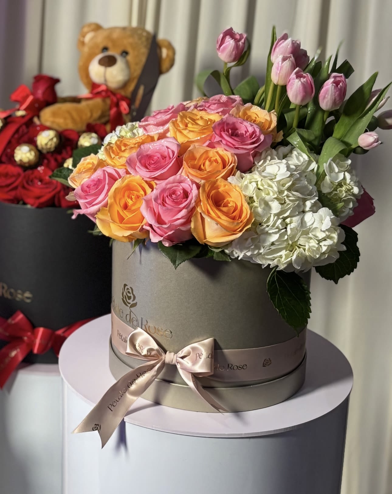 Pink and orange roses with white hydrangeas in a round box, with a teddy bear behind it.