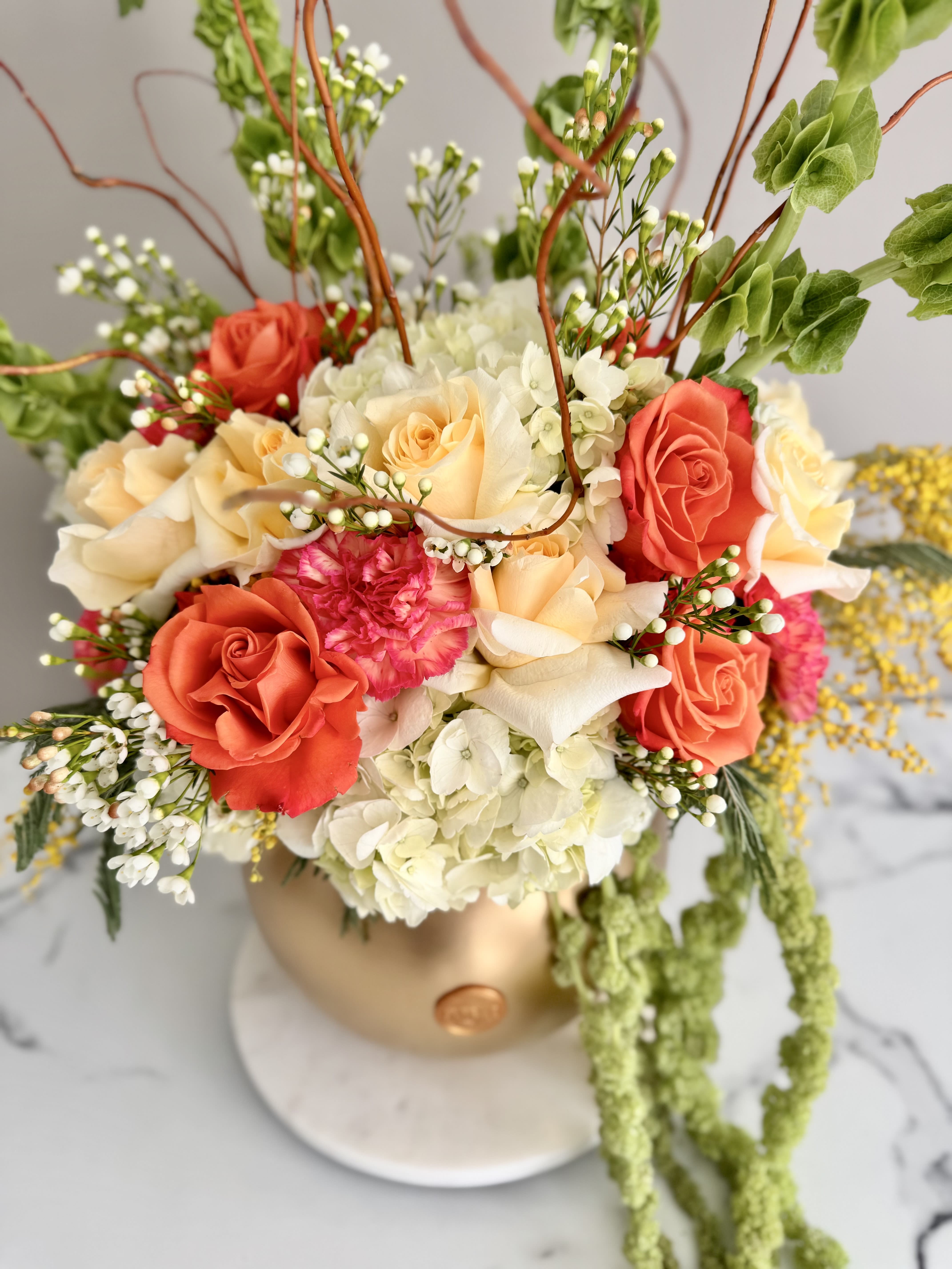 Coral and cream rose arrangement in a gold vase with trailing greenery