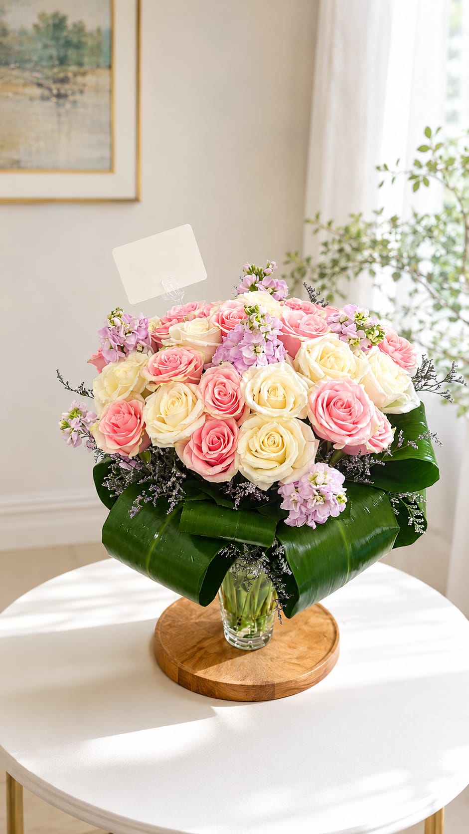 Blushing Elegance - A full cluster of pink and cream roses softened by lavender filler gives this flower arrangement a quietly romantic feel-perfect as a birthday bouquet, anniversary arrangement, or a thoughtful just-because gift that makes someone feel truly seen. The pastel mix of pink, white and purple reads gentle and refined, so the recipient will notice both the color and the care behind it.  Hand-tied and presented in a clear glass vase, the roses are tucked among small lavender blooms and framed by broad glossy leaves wrapped at the base with a decorative leaf wrap for a lush, structured silhouette.  This is the kind of bouquet that brightens an ordinary day and marks special moments with subtle elegance; order standard sizing for a balanced table arrangement or contact us for larger options if you want a bolder statement. To keep the roses fresh, change the water every other day and trim stems slightly when you refresh the vase.