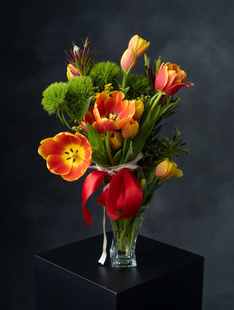 Too Cute Tulip Bouquet - Sunlight-packed tulips in a cheerful spring palette make an instant "she's on my mind" moment - perfect as a birthday bouquet, anniversary surprise, or a bright-thinking-of-you gift. This Too Cute Tulip Bouquet reads playful and affectionate, so it's also a sweet pick for Mother's Day, a housewarming, or to say thank you with a splash of color that feels like a warm hello.  Loose, upright stems of red, peach, orange, yellow and pink tulips are arranged with bright green pom-pom accents and delicate yellow filler blooms in a clear glass vase. A red satin ribbon (plus a touch of white ribbon) is tied around the stems for a gift-ready finish; the arrangement's lively mix of hues and textures creates a casual, energetic silhouette that looks hand-gathered. Ideal for summer and holiday tables alike, it arrives ready to display - a joyful, low-fuss arrangement that will make the person you're celebrating feel genuinely seen.