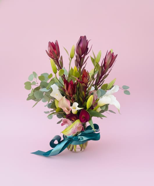 Languid Lilies Vase - Tall, airy calla lilies and lilies create an elegant, contemporary statement-perfect as an anniversary bouquet, a refined birthday gift, or a "just because" surprise for the person you want to make feel treasured. This flower arrangement suits love and romance or more solemn moments like sympathy and memorials: the upright silhouette and mix of soft and jewel tones deliver quiet confidence whether you're celebrating a new baby, saying thank you, or marking Father's Day.  The Languid Lilies Vase pairs white and blush-pink calla lilies with deeper burgundy tropical blooms and protea accents, softened by round eucalyptus foliage for a fresh green contrast. Arranged in a clear glass vase and finished with a wide teal satin ribbon, the palette reads romantic and modern-pink, purple, white, and green layered for texture and depth. Its taller, airy profile gives presence without overwhelming a bedside, mantel, or reception table.  Designed to make the recipient feel truly seen, this arrangement works beautifully for weddings, housewarmings, retirements, get-well wishes, and winter or summer celebrations alike. For best longevity, keep the vase topped up with fresh water and place away from direct heat; available in a single polished size ideal for tabletop display and delivery-ready for the moment you need to say "thinking of you."