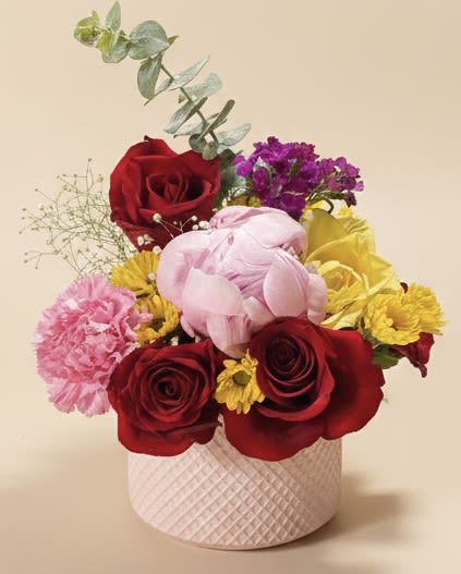 Debutante Hat - A playful mix of deep red roses and soft pink blooms pops against sunny yellow accents and sprightly purple filler flowers-arranged to feel like a modern debutante hat. This flower arrangement for birthdays, anniversaries, or a romantic surprise sits in a pale pink textured hat box that gives the bouquet a polished, gift-ready look; the tall eucalyptus stem rising above the blooms adds airy height and a cool gray-green contrast that keeps the design fresh and contemporary.  The hand-tied composition blends chrysanthemums, carnations, roses, and mixed-season blooms in layered, saturated color-red, pink, purple, yellow, and green-so each viewing angle reveals a new cluster of texture and tone. The round pink box doubles as a keepsake container and frames the bouquet for easy display on a table or desk.  Perfect for saying "I'm thinking of you," "thank you," or "happy birthday," this arrangement reads as both romantic and cheerfully celebratory-ideal for anniversaries or a spontaneous just-because gift. Sized to be noticed without overwhelming a room, it arrives ready to enjoy; trim stems and refresh water when repurposing the flowers from the box into a vase to extend their life.