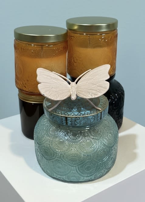  5 Candle Gift Pack - A sculptural small gift set that feels like a mini still life-perfect as a decorative accent or a hostess gift. This flower arrangement alternative pairs three glass jars with metallic lids into a layered vignette that reads modern and thoughtful; suggest it as a birthday gift, housewarming present, or a simple "thinking of you" surprise for someone who loves curated home accents.  The grouping includes two amber-toned jars and one teal patterned jar in front, each with a glossy or textured finish that catches light and adds depth. A delicate white butterfly ornament rests across the front jar to give a soft, whimsical focal point against the blue, orange, black, and white palette. Jars come with metal lids and are sized for tabletop display-compact enough for a mantel, entry table, or bedside dresser.  Give this to the person you want to make feel noticed and cared for: it reads like a little moment of everyday beauty they can place wherever they want. Easy to style and low-maintenance, the set arrives ready to display and makes a polished, giftable alternative when flowers aren't the right fit.