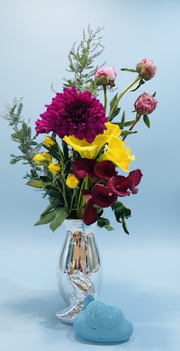 Baby Moon Bouquet - A joyful burst of color that feels both modern and tender - this tall floral arrangement is perfect as a New Baby gift, Thinking of You surprise, or get-well pick-me-up. Bold magenta chrysanthemums and bright yellow calla lilies create an uplifting focal point, while deep burgundy calla lilies and soft pink tulip buds add depth and gentle sweetness. The overall vertical, airy silhouette gives the bouquet a fresh, contemporary presence that makes the recipient feel seen and celebrated.  Arranged in a reflective silver vase and finished with a small blue ceramic cloud figurine at the base, the composition mixes playful charm with polished style. Branching greenery and an open, loose shape let each bloom breathe, offering visual texture and movement from every angle. Sized to stand out on a bedside table or entry console, this bouquet arrives ready to display; light daily water refreshes and a cool spot away from direct heat will help the stems last longer.