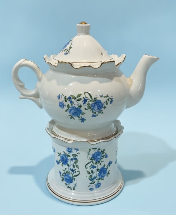 Blue Roses Vintage Tea Pot gift - Delicate blue rose motifs dance across a vintage-style white ceramic teapot-an instantly charming flower arrangement perfect for a "just because" surprise or a heartfelt housewarming gift. The Blue Roses Vintage Tea Pot gift pairs romantic blue and crisp white blooms with soft green accents to create a soothing, thoughtful presentation that feels like a keepsake; it's an excellent choice for thank yous, thinking of you moments, birthdays, retirements, or to bring comfort during get well and sympathy occasions.  The arrangement sits in a gloss white teapot with a matching ceramic stand, both decorated with hand-painted blue roses and tiny green leaves and finished with subtle gold trim around the lid and base. Rounded body, curved handle, and angled spout lend a quaint, collectible look-this piece reads as decorative and homey as much as floral. The mix includes garden-style roses and spray roses in coordinated blue, white, and green tones for a soft, textured bouquet that complements the vintage teapot without overpowering it.  Ideal for a new baby gift set, Mother's Day surprise, or a restful bedside display during recovery, this keepsake teapot makes the recipient feel genuinely seen and cared for. Because the teapot is presented as a decorative vessel, it works equally well displayed on a mantel, kitchen shelf, or dining table; standard floral care-fresh water and trimming stems-will help keep the arrangement looking its best.