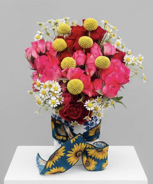 Chamomile Rose - A lively cluster of bright pink and red roses framed by sunny yellow button chrysanthemums and petite white daisies greets the eye-perfect for a birthday bouquet, a "just because" surprise, or a springtime thank-you arrangement. The Chamomile Rose arrangement reads as cheerful and handcrafted, the kind of floral gift that makes the person you're thinking of feel genuinely celebrated, seen, and adored on occasions from anniversaries and graduations to housewarmings and retirements.  Flowers are packed tightly into a rounded, full composition that feels generous and joyful; the roses bring romantic depth while the button mums and daisy-like blooms add playful texture and a sunlit color story. Delivered in a patterned ceramic vase and finished with a wide sunflower-print ribbon, this piece arrives ready to display-no extra styling needed. It's equally suited for weddings, Valentine's Day, Mother's Day, or a casual "thinking of you" moment; for best results, keep the vase water fresh and trim stems every few days to extend the life of this bright, heartwarming arrangement.