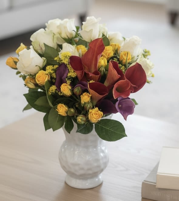 Coffee table Vase - A bright, jewel-toned centerpiece that makes someone feel celebrated the moment it arrives-this coffee table vase arrangement blends white roses, sunny yellow spray roses, and bold red and purple calla lilies for a lively, elegant statement perfect for a birthday, housewarming, or "just because" surprise. The mix of soft white blooms and saturated hues reads both cheerful and refined, so it also suits thank you gestures, thinking-of-you moments, and romantic occasions like anniversaries or Valentine's Day.  Arranged in a low, rounded clear glass vase, the compact shape stays full and balanced-ideal for coffee tables, entry consoles, or bedside dressers. Texturally rich spray roses nestle around classic roses while the sculptural calla lilies add height and drama without overwhelming the design. The polished glass keeps the focus on the flowers and their mixed color palette, making this a versatile choice for spring and summer gifting, new baby celebrations, graduations, or to brighten a get-well room.  This arrangement is designed to make the recipient feel genuinely seen-whether you're saying thank you, offering sympathy, celebrating a milestone, or sending love. Easy to place and low enough for conversation-friendly tabletops, it arrives ready to enjoy; simple care-fresh water and a cool spot-will keep the blooms looking their best through the week.