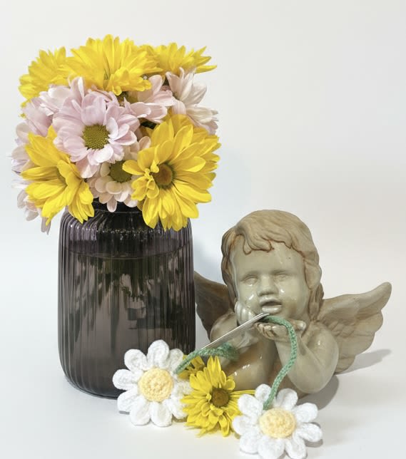 Daisy Jar w/crocheted daisies - A burst of sunny daisies in bright yellow, soft pink and crisp white brings an effortless, cheerful note to any "just because" moment-perfect as a birthday surprise, a thank-you gift, or a lighthearted anniversary bouquet. This daisy jar arrangement reads like spring in a vase: playful, fresh, and quietly joyful, the sort of present that makes the person you're thinking of feel genuinely seen and smiling.  Clusters of daisy-style blooms are gathered in a dark ribbed glass vase that adds vintage texture and contrast to the pale tones. A daisy garland is draped in front and a small cherub figurine sits beside the vase for a whimsical, decorative touch that complements mantle, bedside table, or kitchen windowsill displays. The compact composition fits well for everyday gifting or seasonal spring décor without overwhelming a room.  Ideal for saying "thank you," celebrating a birthday, marking an anniversary, or surprising Mom on Mother's Day, this arrangement offers an approachable, long-lasting look that suits multiple occasions-including Valentine's Day when you want something sweet and not traditional red. Care: keep blooms in fresh water, out of direct midday sun, and refresh water every other day to keep the daisies perky.