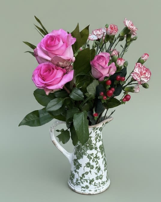 Elegant Vintage Bouquet w/ keepsake pitcher vase - Two soft pink roses take center stage in this floral arrangement, making it an ideal birthday bouquet, anniversary gift, or just-because surprise to let someone know you're thinking of them. Designed for occasions from a heartfelt thank-you to a housewarming or bridal shower, the Elegant Vintage Bouquet reads as gentle and romantic-perfect for Mother's Day, Women's Day, or to say "I love you" without being over the top.  Clusters of pink-and-white carnations and delicate stock surround the roses, while dark green foliage and a few red berry accents add contrast and texture. The stems are gathered in a white ceramic pitcher patterned with green florals, giving the design a cottage-style, vintage charm that doubles as a keepsake vase. The color story of pink, white, green, and touches of red feels fresh for spring and summer but remains timeless enough for any season.  This arrangement suits the person you want to make feel truly seen-whether it's a best friend, grandparent, or the new homeowner. The ceramic pitcher arrives ready to display; light daily watering and a fresh trim to the stems every few days will keep the blooms looking their best.