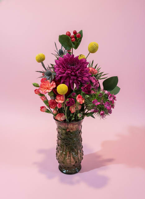 Exotic Intoxica - A compact, upright spray of vivid magenta, coral, yellow and purple blooms brings instant cheer-perfect as a just-because bouquet or a colorful housewarming, birthday, or get-well gift. The arrangement's modern, slightly whimsical silhouette makes the person you're thinking of feel truly seen: bold color, playful texture, and a confident focal bloom that reads like a happy exclamation.  A large purple chrysanthemum anchors the center surrounded by coral and magenta carnations, yellow button-style chrysanthemums, protea accents and other mixed blooms for a sculptural, clustered look. Arranged in a textured dark glass vase, the composition is intentionally compact and easy to place on a bedside table, entry console, or desk. Its bright mixed colors suit thank-you, thinking-of-you, I'm-sorry, anniversary, retirement, or congrats moments and work beautifully year-round.  Size and care: vase-arranged and ready to display; swap water every couple days, trim stems if removed from the vase, and keep out of direct heat or sunlight to extend freshness.