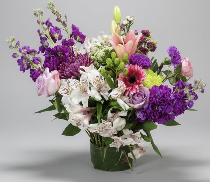 Fairy Garden favorites - A bright, garden-style bouquet that feels like a secret patch of spring, perfect as a thank-you or thinking-of-you gift. This low glass vase arrangement is built around tall lily buds and branching purple accents that rise above a lively mix of pink, lavender, white and green blooms-an everyday floral gift that also suits birthdays, new baby celebrations, or a cheerful just-because surprise.  The mix includes lilies, alstroemeria, chrysanthemums, freesia, roses and a sunny gerbera daisy arranged with textured greenery for depth and movement. The layered composition pairs stronger purples with soft blush and white tones so each flower reads clearly: tall buds and sprays add vertical interest while the fuller roses and mums fill the base for a generous, full look. The clear glass vase keeps the focus on the blooms and works well on a table, bedside dresser, or office desk.  Send it to make someone feel genuinely seen-this is the kind of bouquet that brightens a new-parent room, marks a spring birthday, or says thank you without fanfare. The arrangement arrives ready to display; keep water fresh and trim stems occasionally to extend vase life.