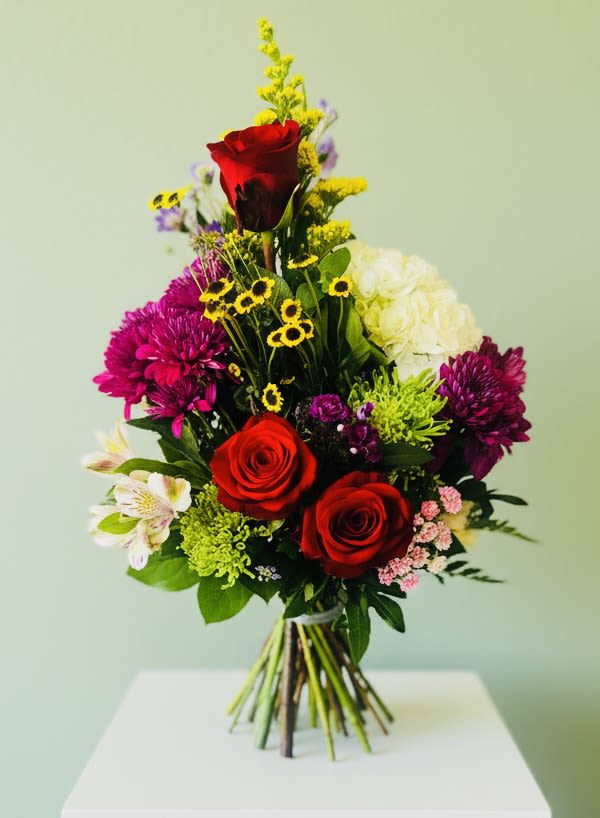 Floral Drama - A bold, upward-reaching bouquet that reads like a celebration in bloom - Floral Drama is a tall mixed arrangement perfect for a birthday, anniversary, or a spontaneous "just because" that says love and appreciation. Framed by two prominent red roses and a vertical line of blooms, the bouquet delivers an immediate romantic impression while the mix of colors keeps the mood joyful rather than formal.  This hand-tied design layers red roses with pink chrysanthemums, crisp white blossoms, purple accents, and bright yellow filler for contrast, creating a dense, textured composition with a full, rounded base and a lively, vertical silhouette. The palette of reds, pinks, purples, whites, and greens gives the recipient a rich sensory experience - visual warmth, soft petal textures, and an abundant, fresh look that suits both romantic gestures and heartfelt thank-you moments.  Ideal for the person you want to make feel seen and celebrated - whether marking an anniversary, saying "I love you," or surprising someone on their birthday - Floral Drama arrives as a substantial, eye-catching bouquet that reads special in any season. For best enjoyment, trim stems and refresh water daily to keep the mixed blooms vibrant.