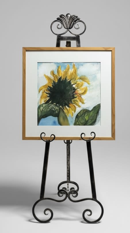 Framed Watercolor of a sunflower by local artist - A single sunflower painted in warm, honeyed yellow takes center stage in this framed watercolor-an inviting home accent that feels like a bright, personal note for housewarming, birthday, or just because. The sunflower artwork reads as a thoughtful gift for Father's Day, Mother's Day, or an anniversary when you want the person you're thinking of to feel seen; its sunny presence also works beautifully for spring and summer decor or a wedding welcome table.  Rendered on canvas with soft blue-and-white washes behind broad green leaves, the piece is set in a wood-toned frame with a metallic gold outer edge and finished with a canvas backing and corner supports for durability. It arrives ready-to-display on the included ornate black metal easel with curled details, so the recipient can place it on a mantel, entry table, or shelf without additional hardware.  This framed sunflower is a simple, enduring way to say thank you, congrats, or thinking of you-perfect for best friends, new homeowners, or anyone who loves cheerful, rustic art. Size and framing details are included on the label and canvas backing; care is minimal-keep out of direct sunlight and dust gently-so it stays a sunny focal point for years.